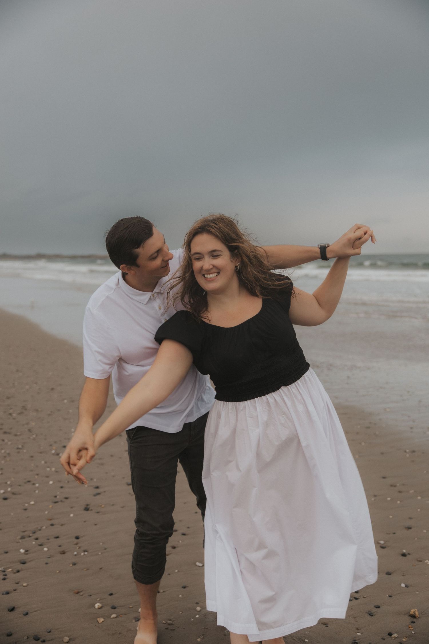 A joyful couple dancing on a sandy beach with ocean waves and cloudy skies behind them.