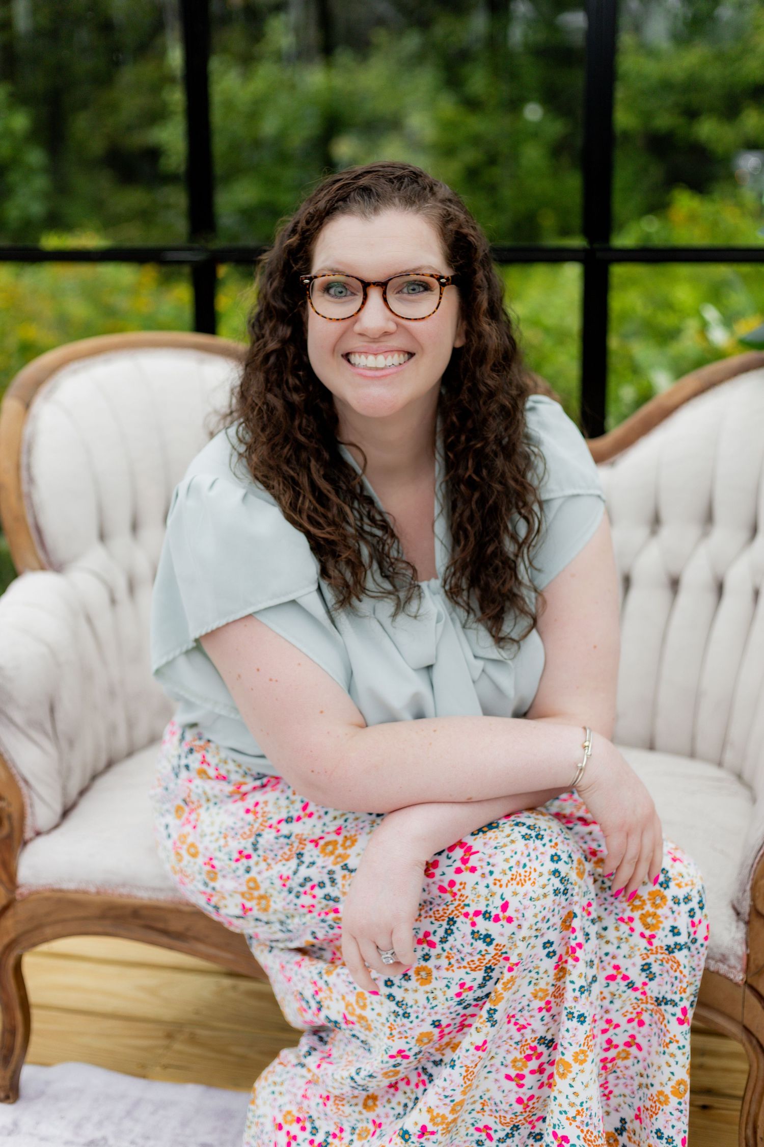 Person wearing a light blue top and floral skirt relaxes on a tufted vintage sofa outdoors.
