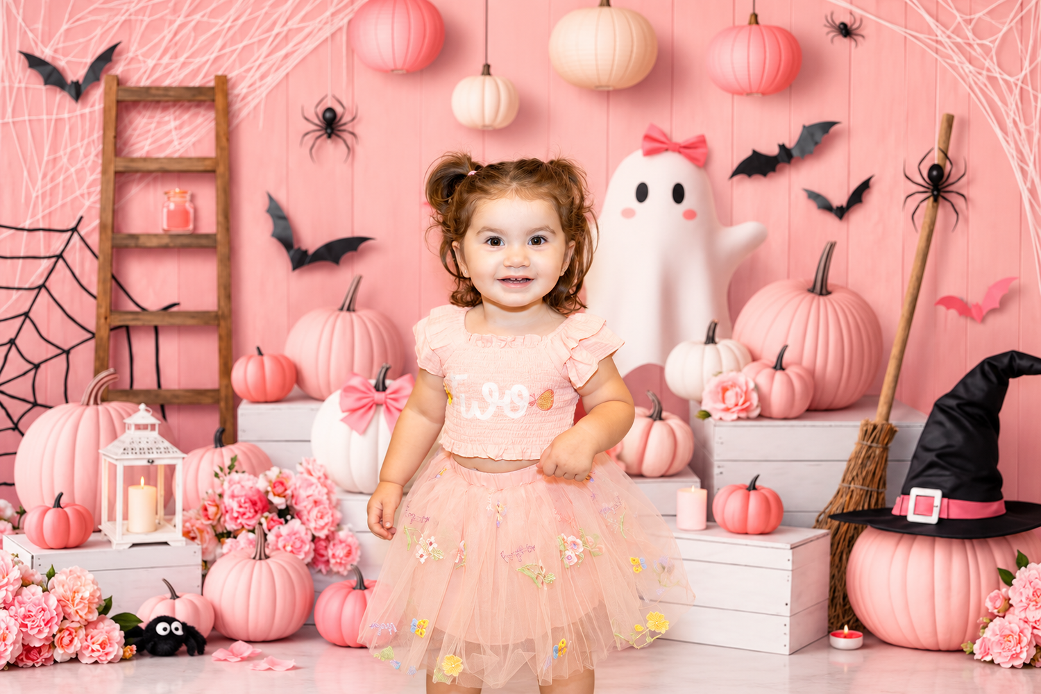 A toddler girl in a pink tutu poses in a Halloween-themed pink set with pumpkins, ghost, bats, and witch decorations.