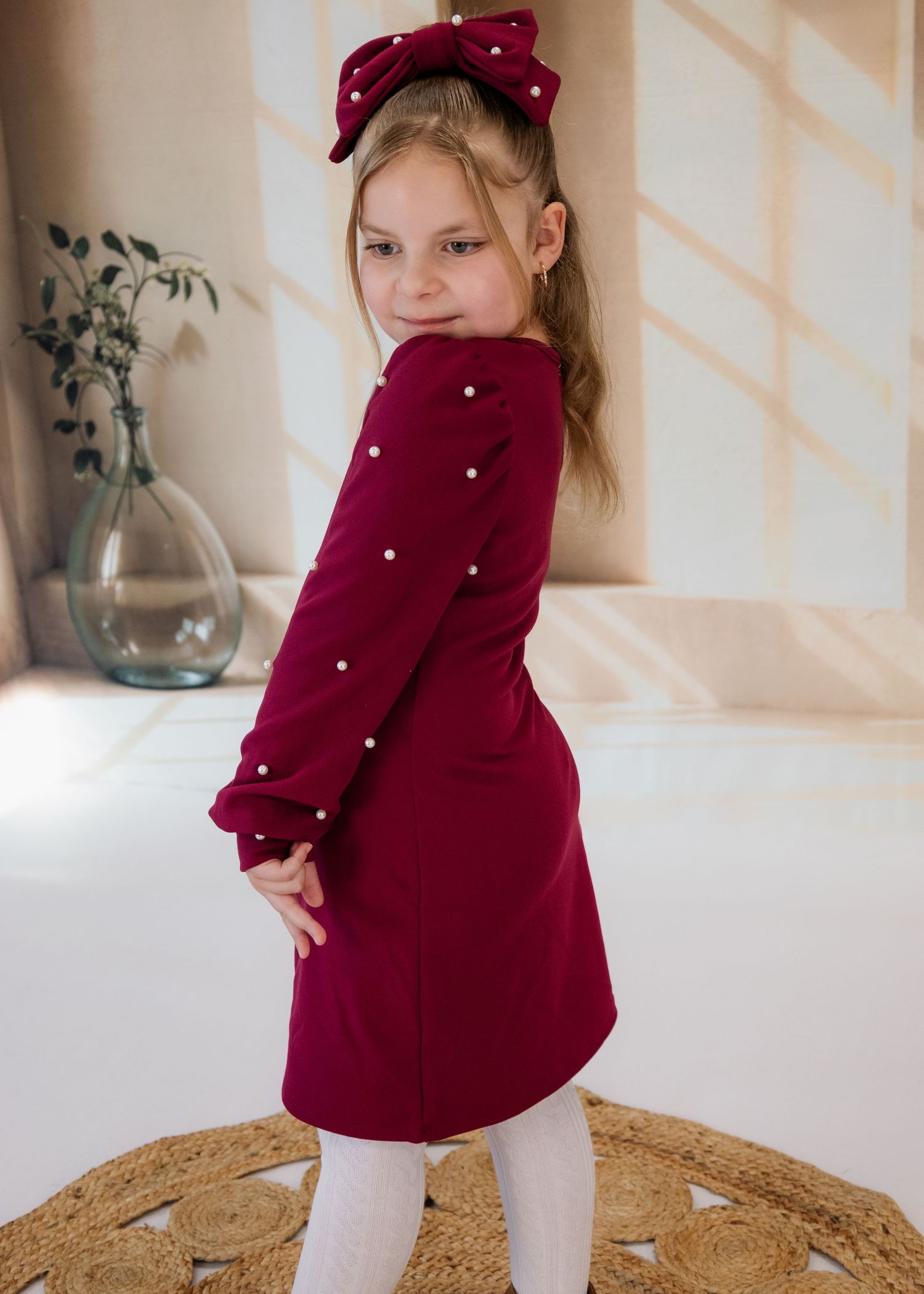Small child in burgundy dress twirling on round jute rug in bright room.