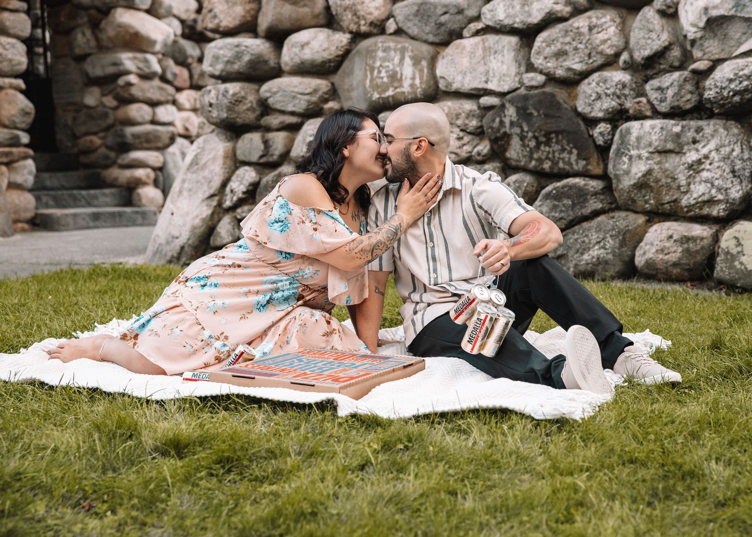 Couple sharing a romantic kiss while sitting on a blanket in the grass against a rustic stone wall backdrop.