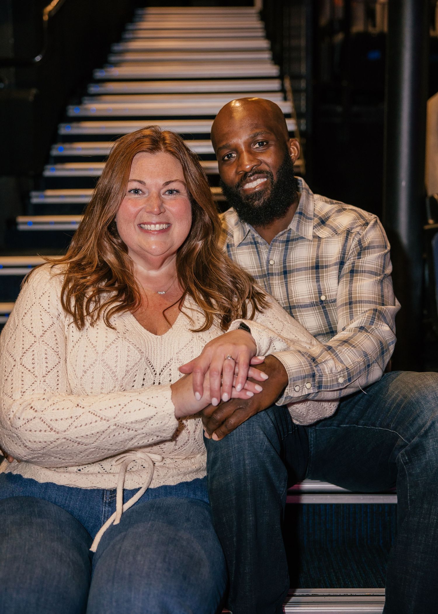 Smiling woman in a cream knit sweater and bearded man in a plaid shirt sit close together holding hands on indoor stairs.