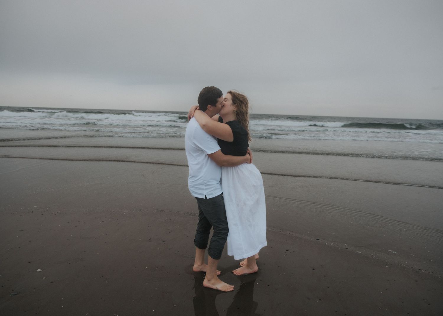 Couple embracing and kissing barefoot on a wet sandy beach with gentle waves and a cloudy gray sky in the background.