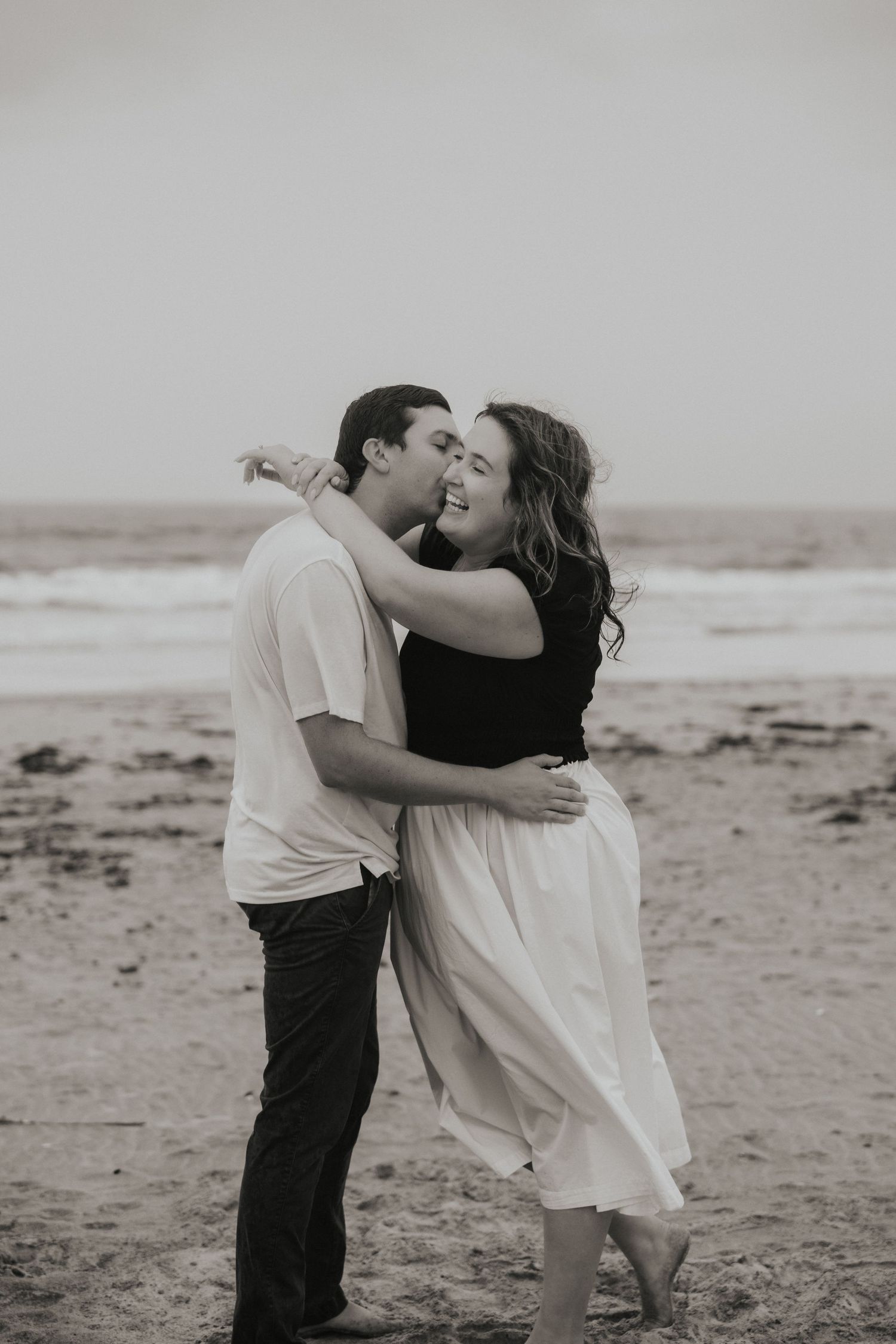A couple embraces and laughs together on a sandy beach in a romantic black and white photo.