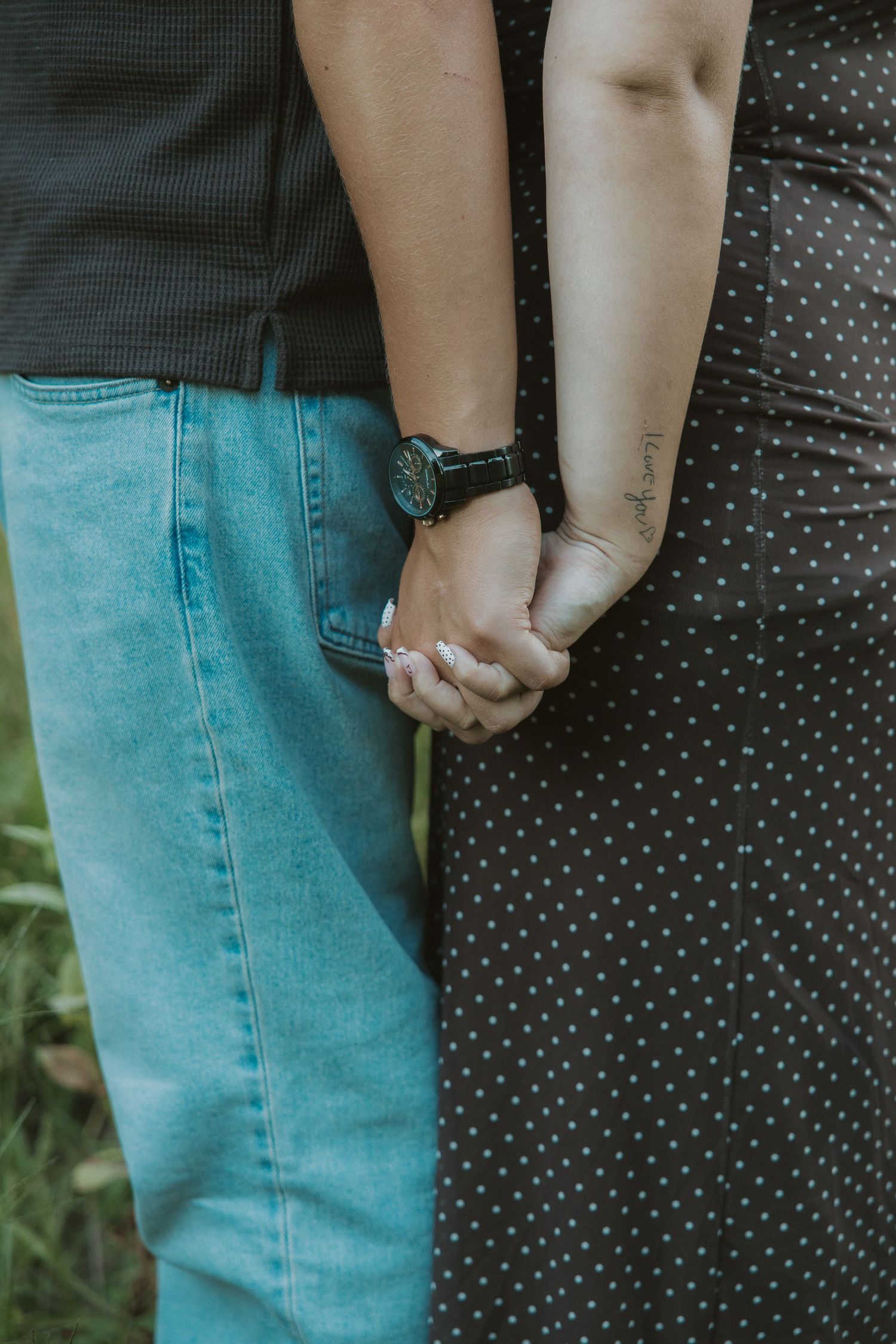 Close-up of a couple holding hands, one wearing a watch and the other with a forearm tattoo, in casual outdoor setting.