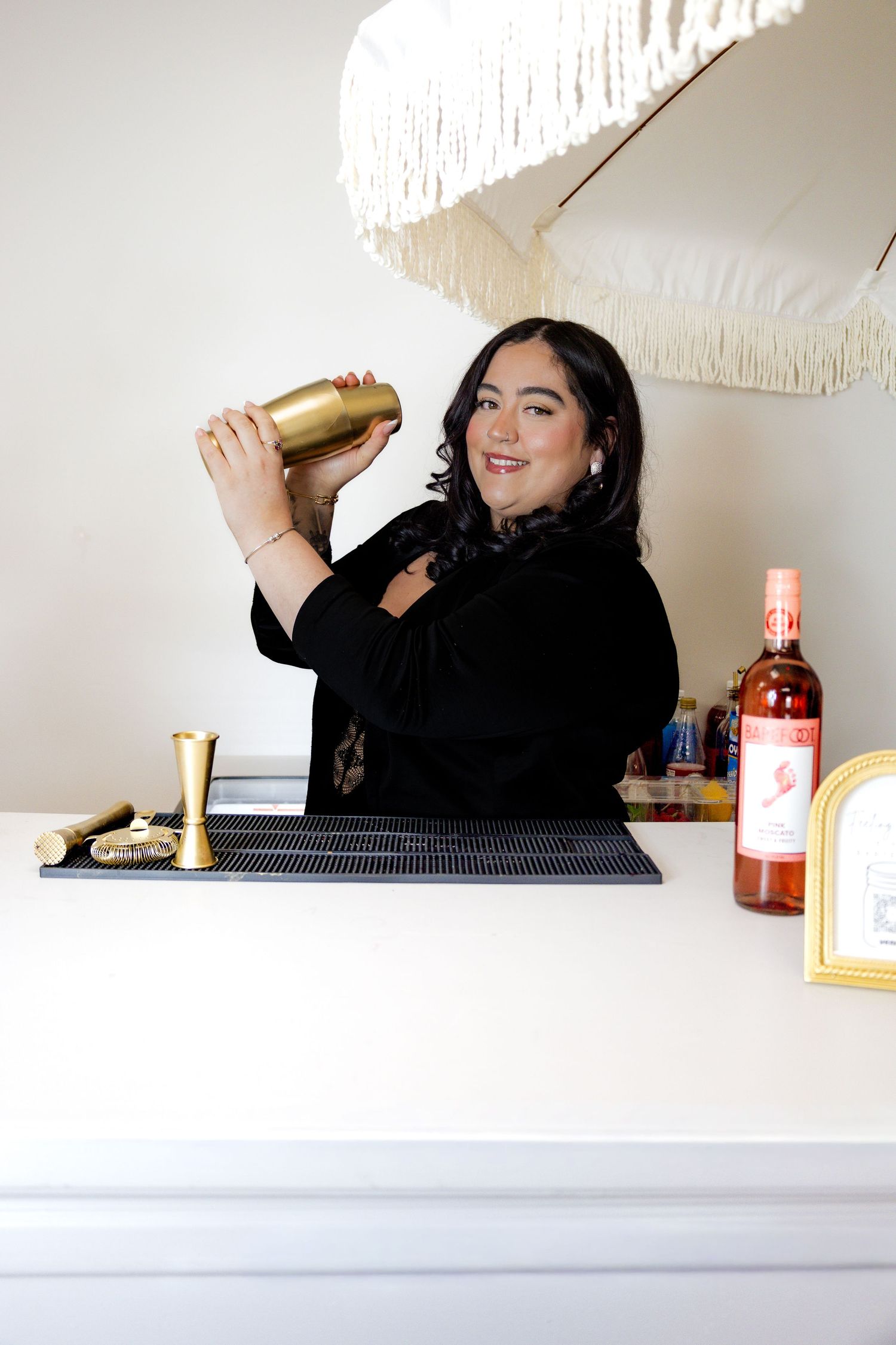 Woman in black dress smiling while shaking gold cocktail shaker behind a white bar.