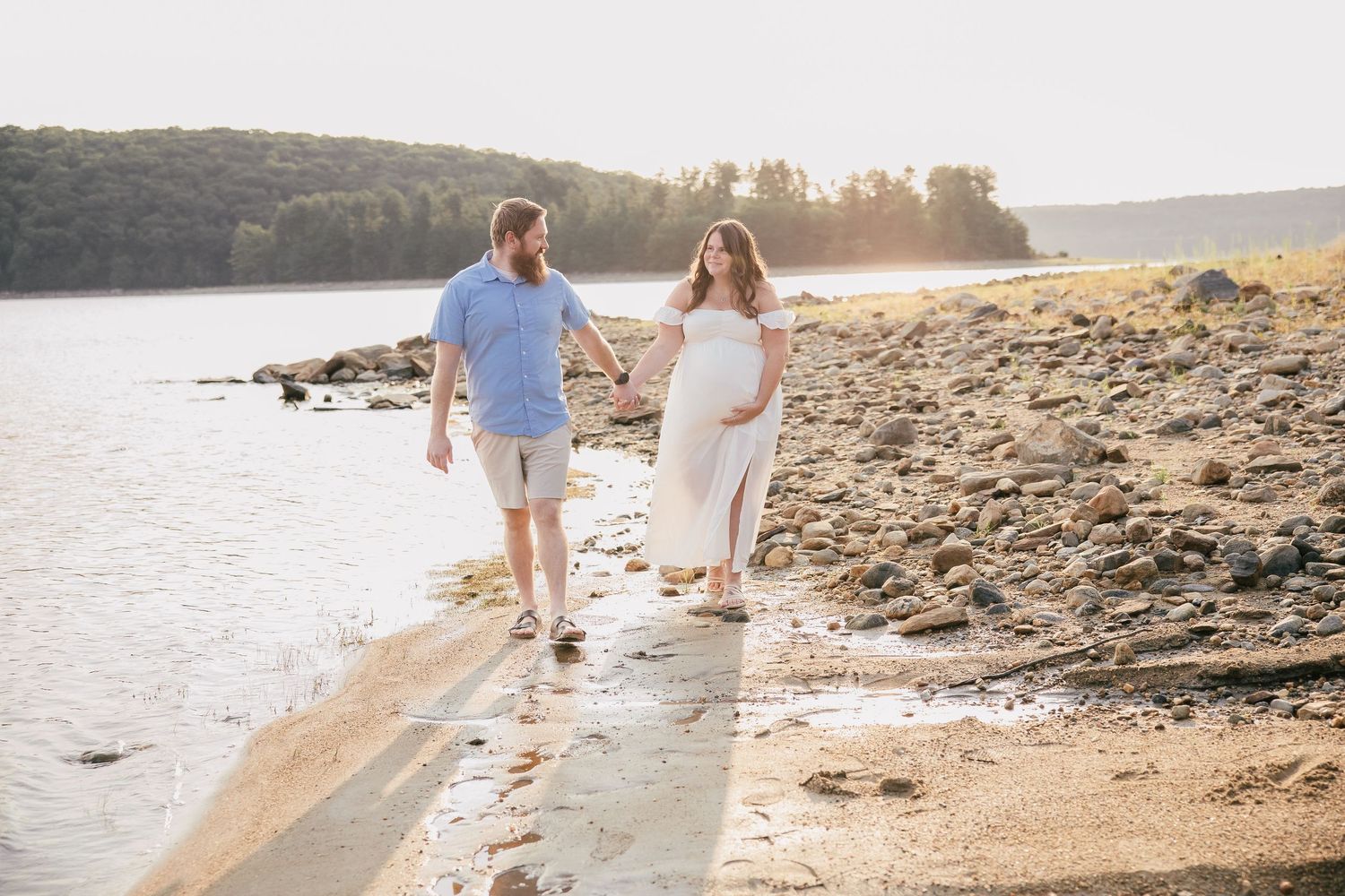Pregnant couple walking hand-in-hand along a sunlit rocky shoreline, woman in white dress, man in blue shirt.