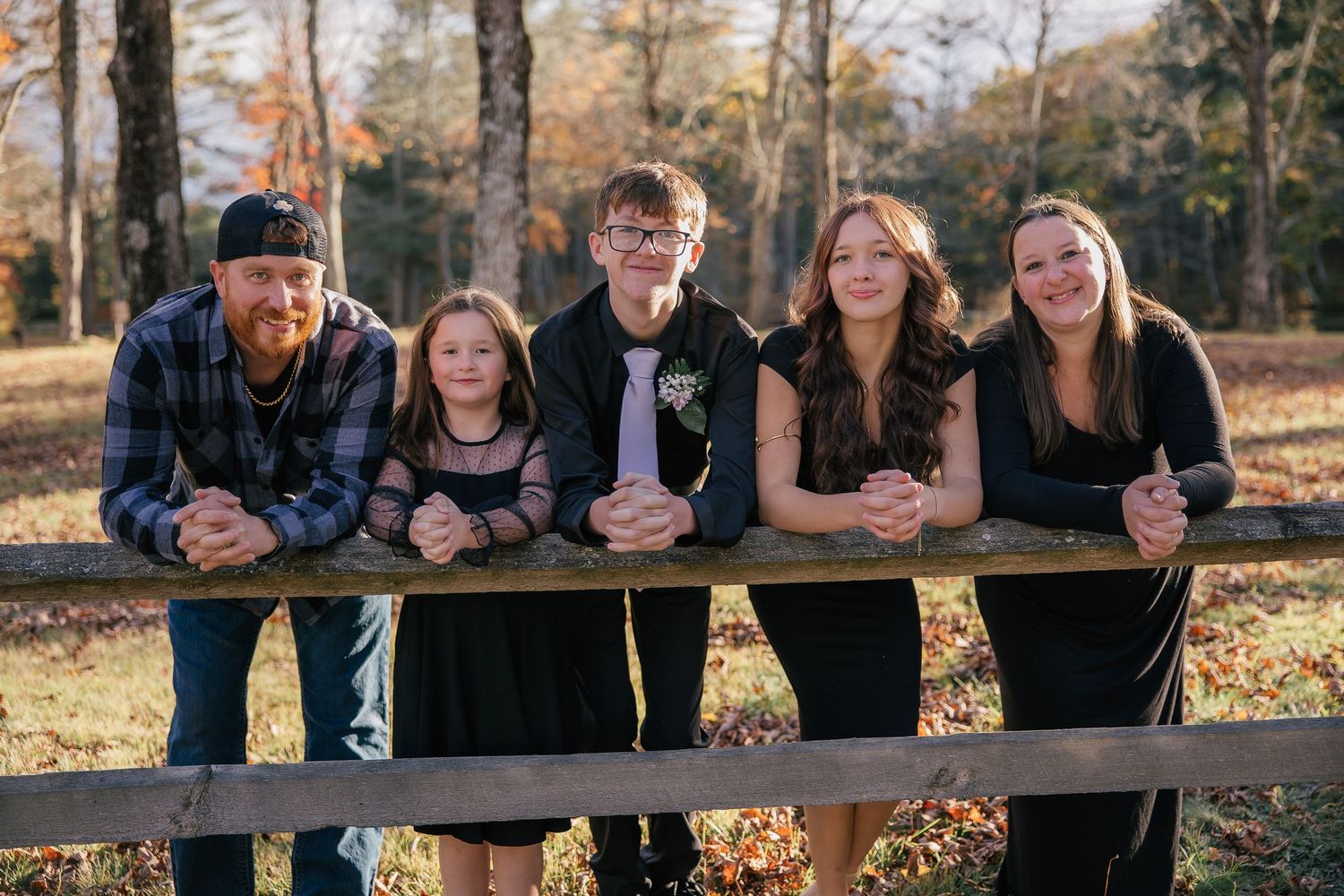 Family of five posing along a wooden rail fence in an autumn woodland setting, smiling together during a outdoor portrait session.