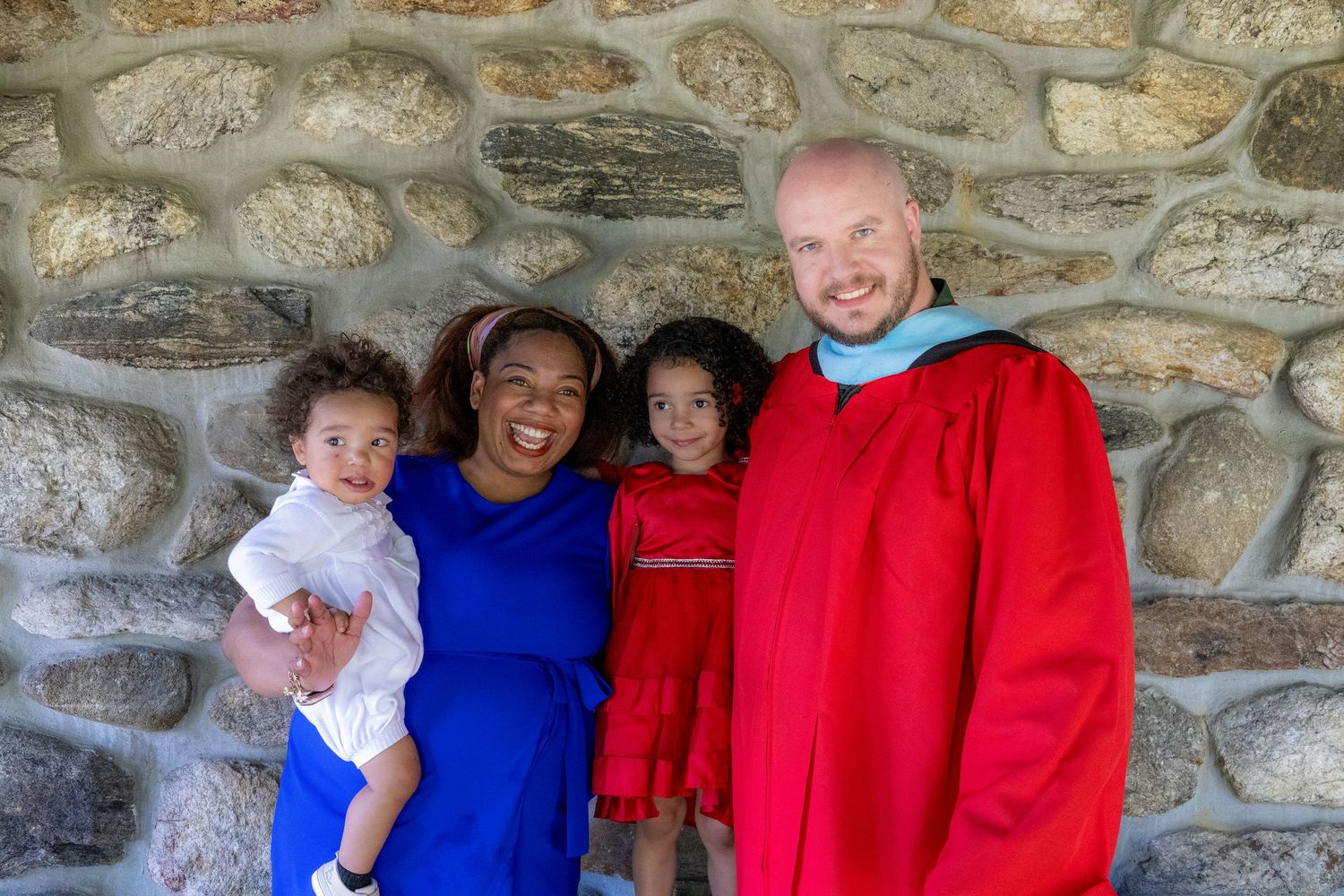 Smiling graduate in red robe poses with family of four against rustic stone wall background.