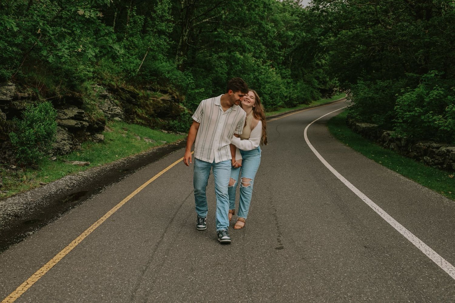 A couple walks hand in hand down a winding forest path surrounded by lush green trees.