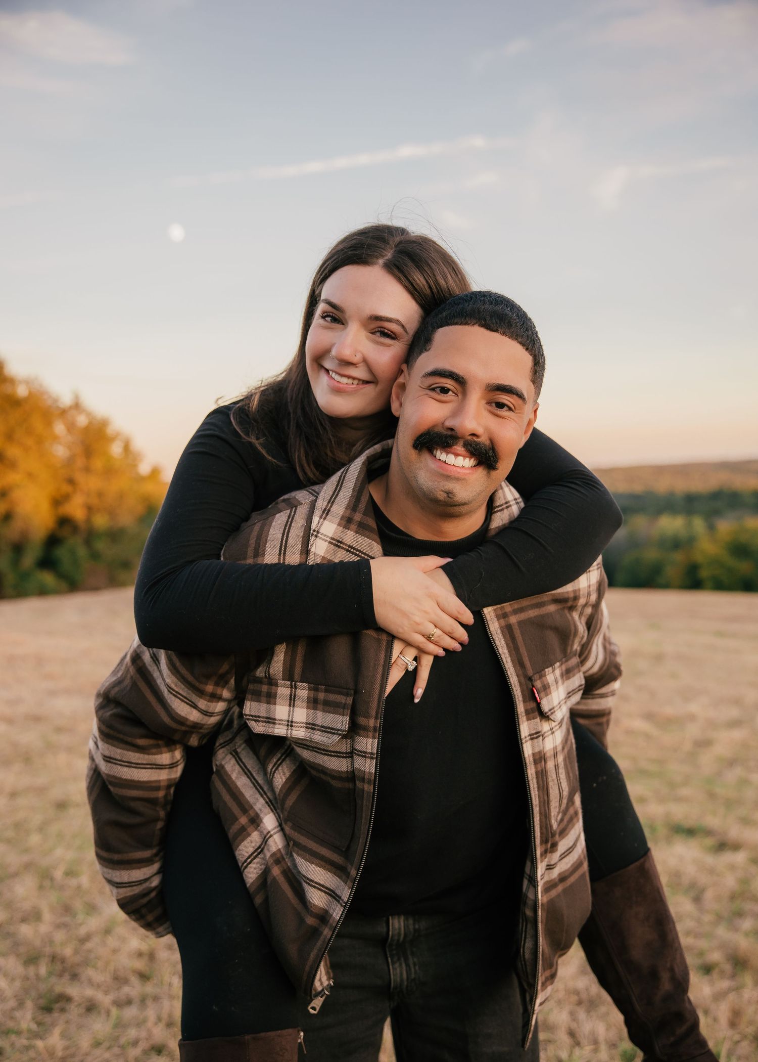 A smiling couple poses outdoors at golden hour, woman hugging man from behind in a field with autumn foliage.