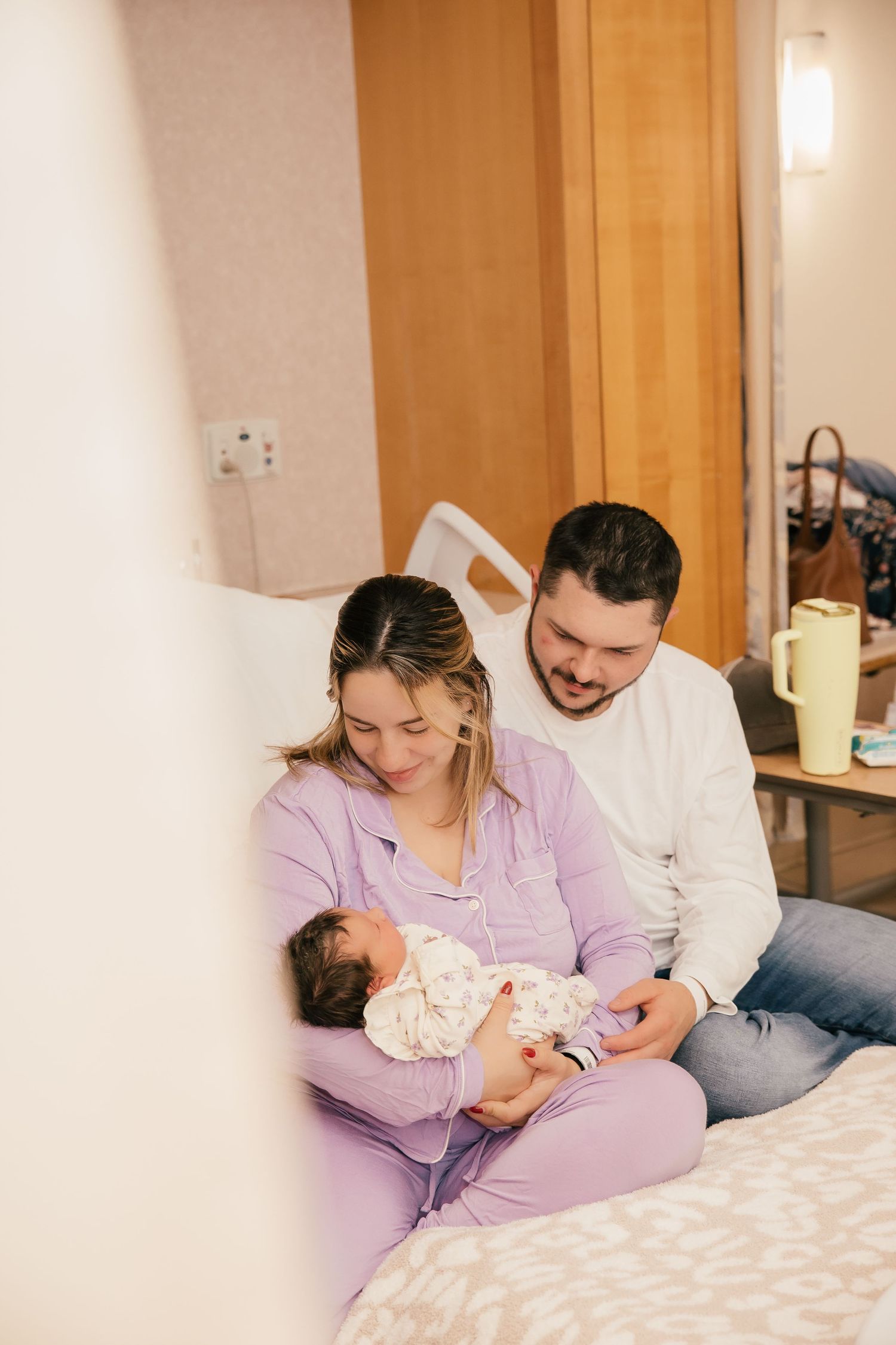New parents sitting on hospital bed smiling at their newborn baby wrapped in a floral blanket.