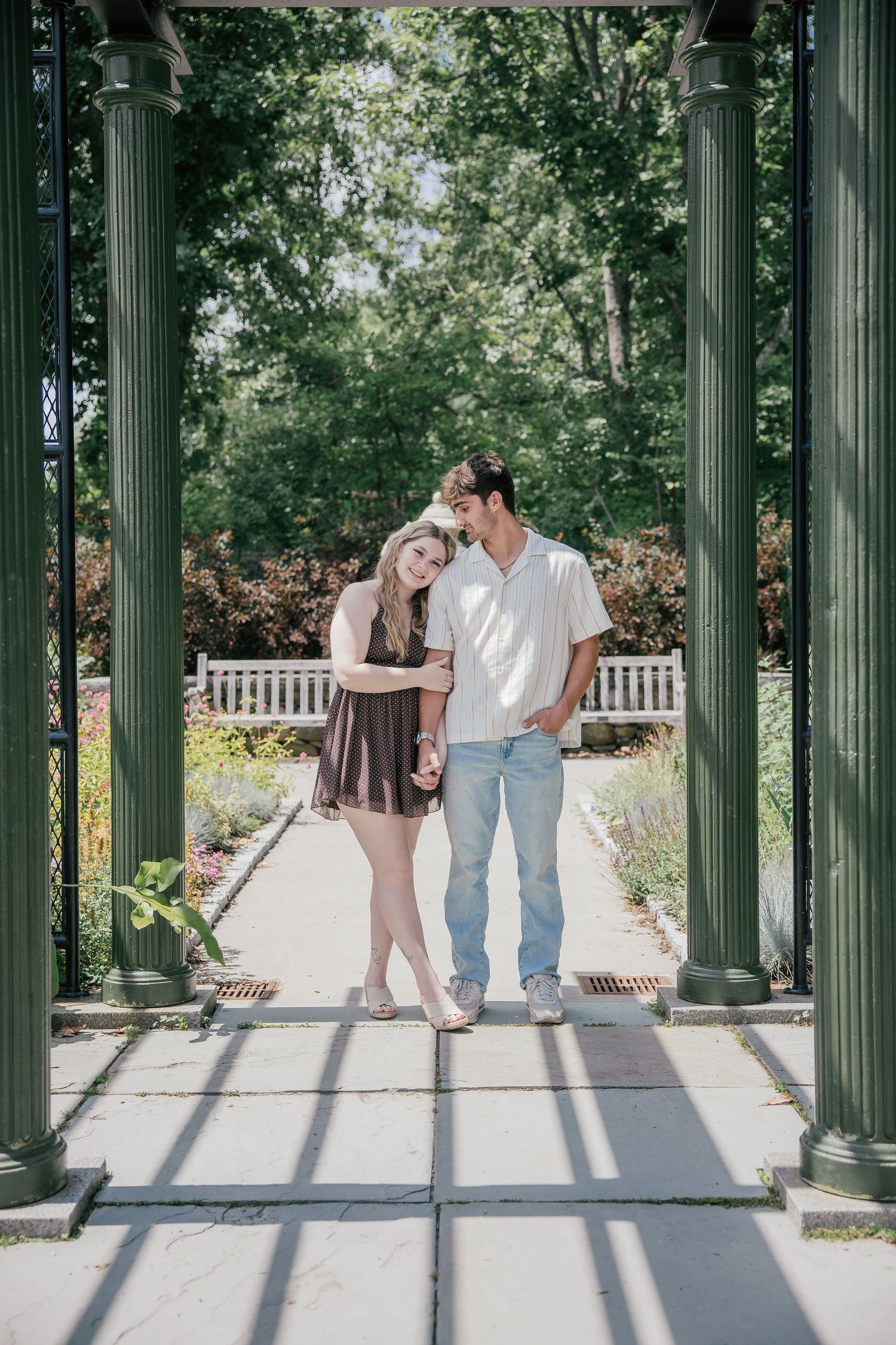 Couple posing under green columns in a garden gazebo, woman in brown dress, man in white shirt and jeans.