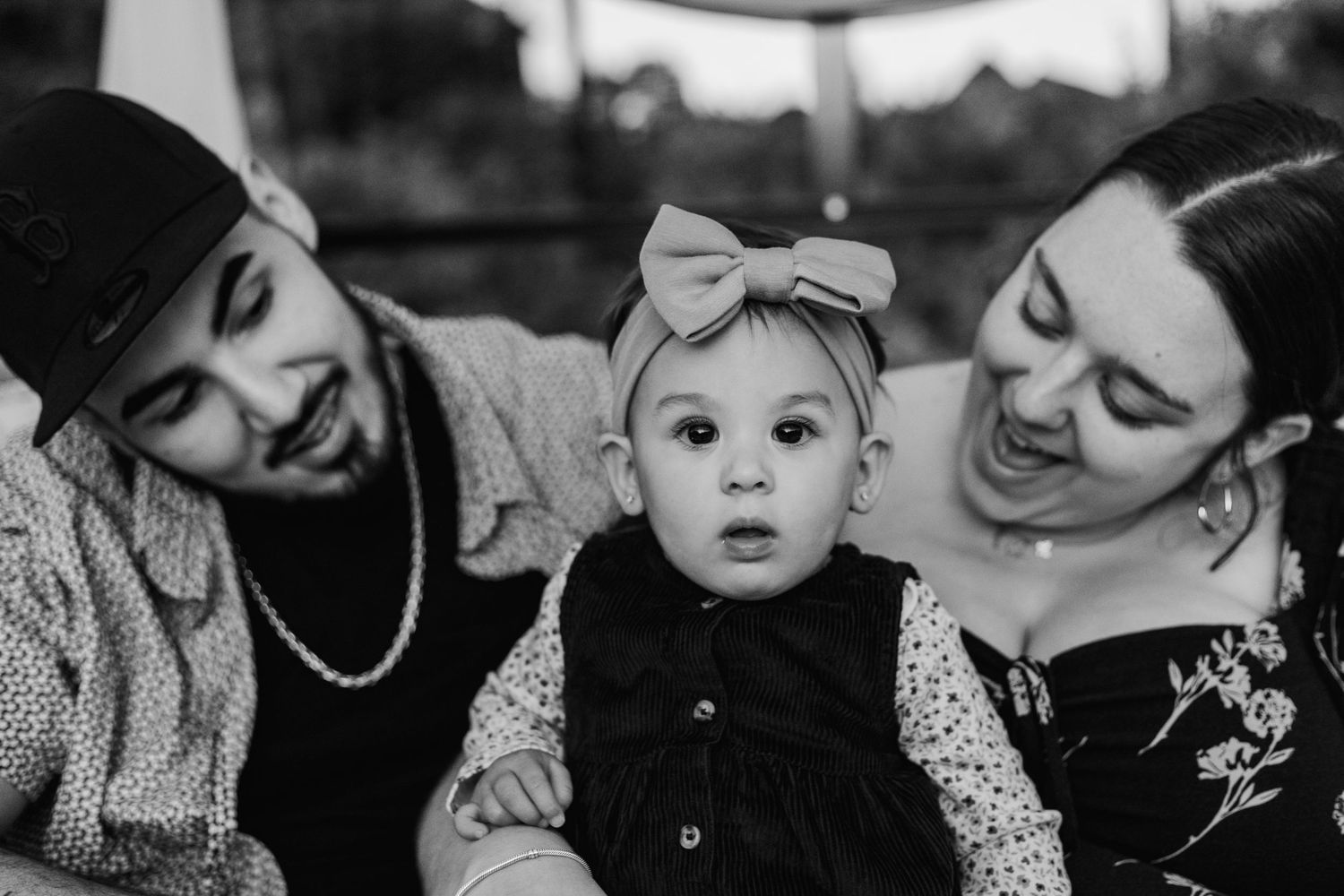 A black and white portrait of a baby with a bow headband between two adults.