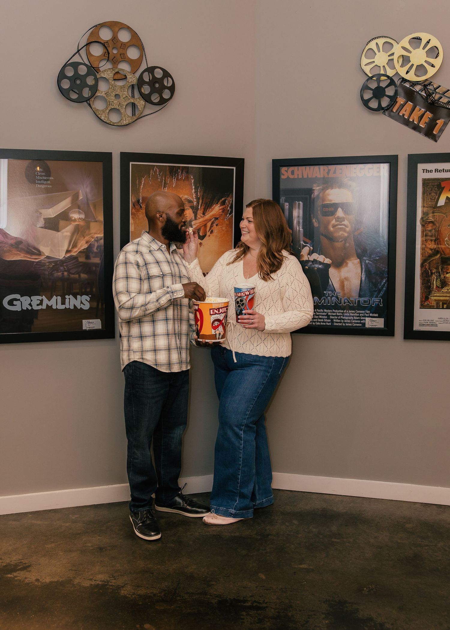 Couple sharing popcorn in front of classic movie posters including Gremlins and Terminator with film reel decorations.
