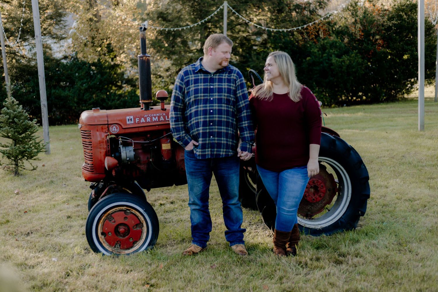 Couple posing together in casual attire next to a vintage red tractor in a countryside setting.