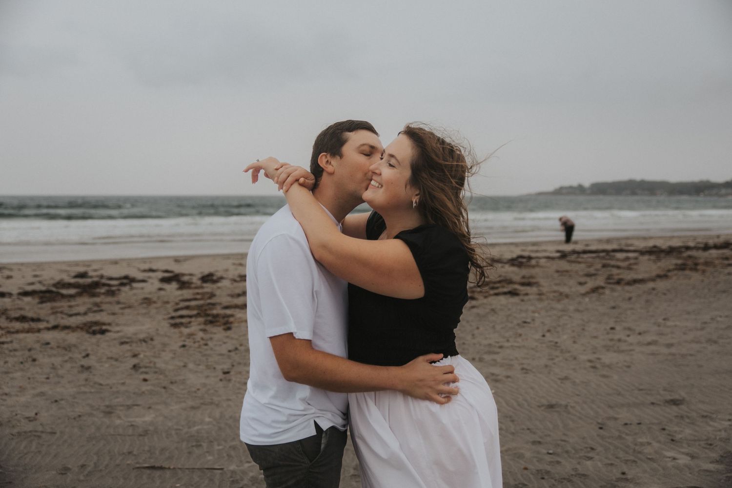 Couple embracing and kissing on a sandy beach with wind-blown hair, calm ocean and overcast sky behind them.