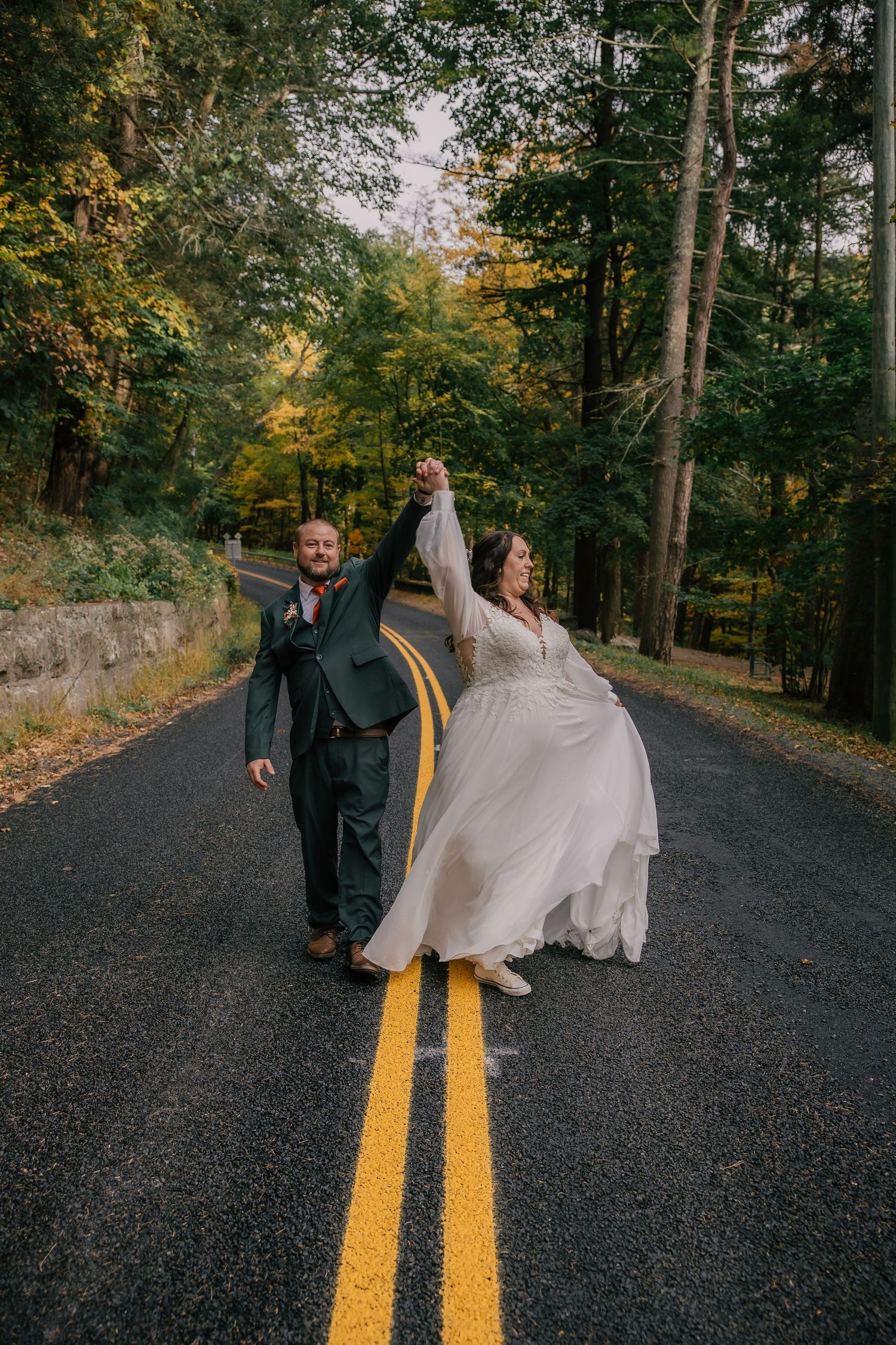 Newlywed couple walks down tree-lined road holding hands raised in celebration during fall wedding.