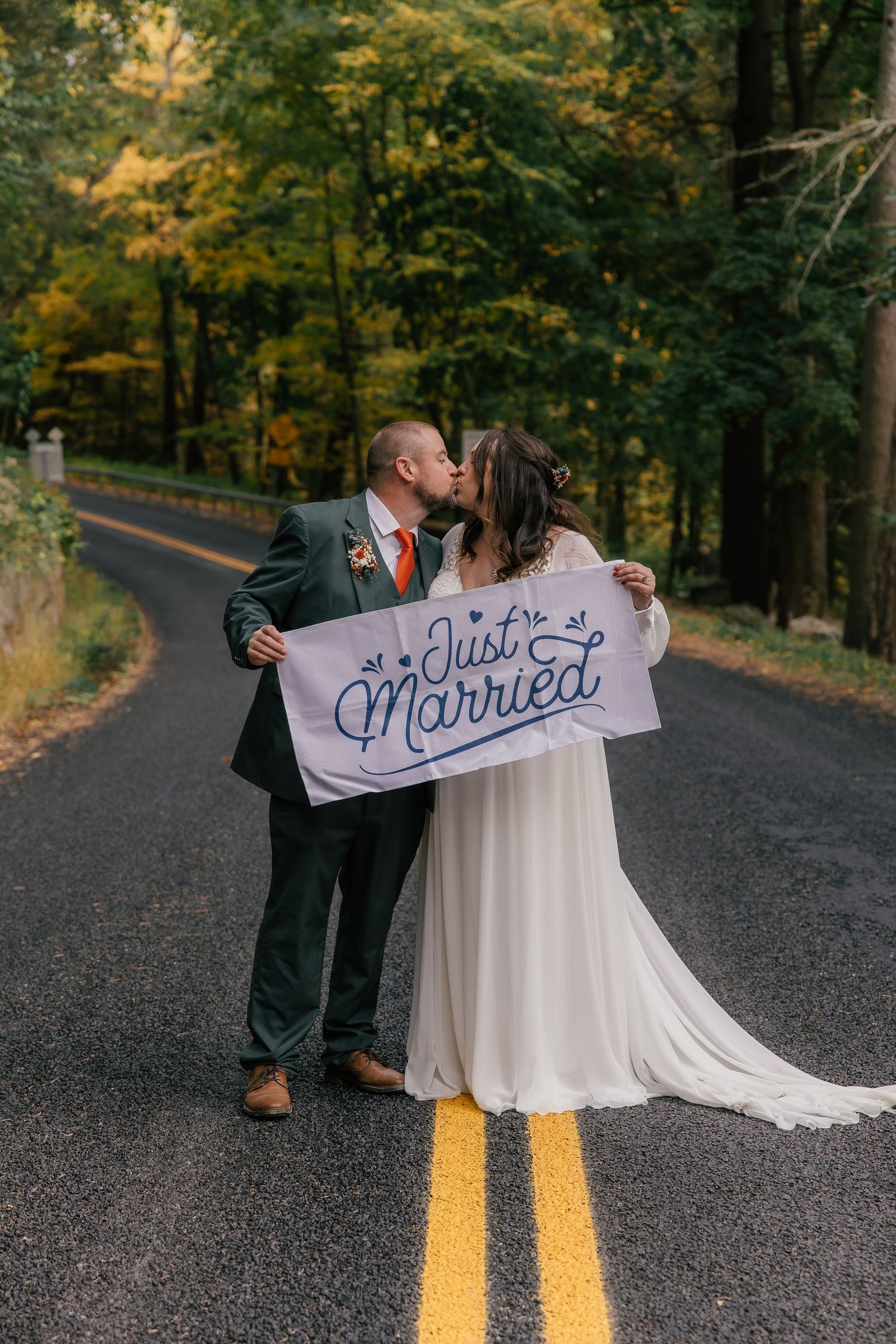 Newlywed couple kissing on a tree-lined road holding a Just Married sign in autumn.