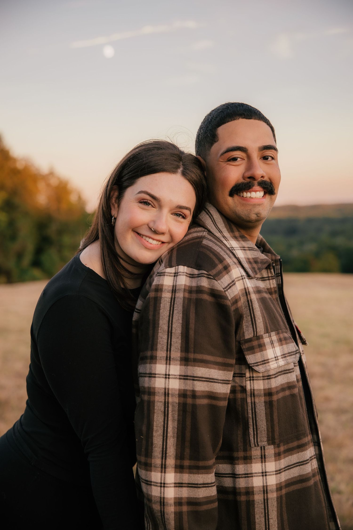 A smiling couple poses at golden hour in a field, woman in black top leaning on man in plaid shirt.