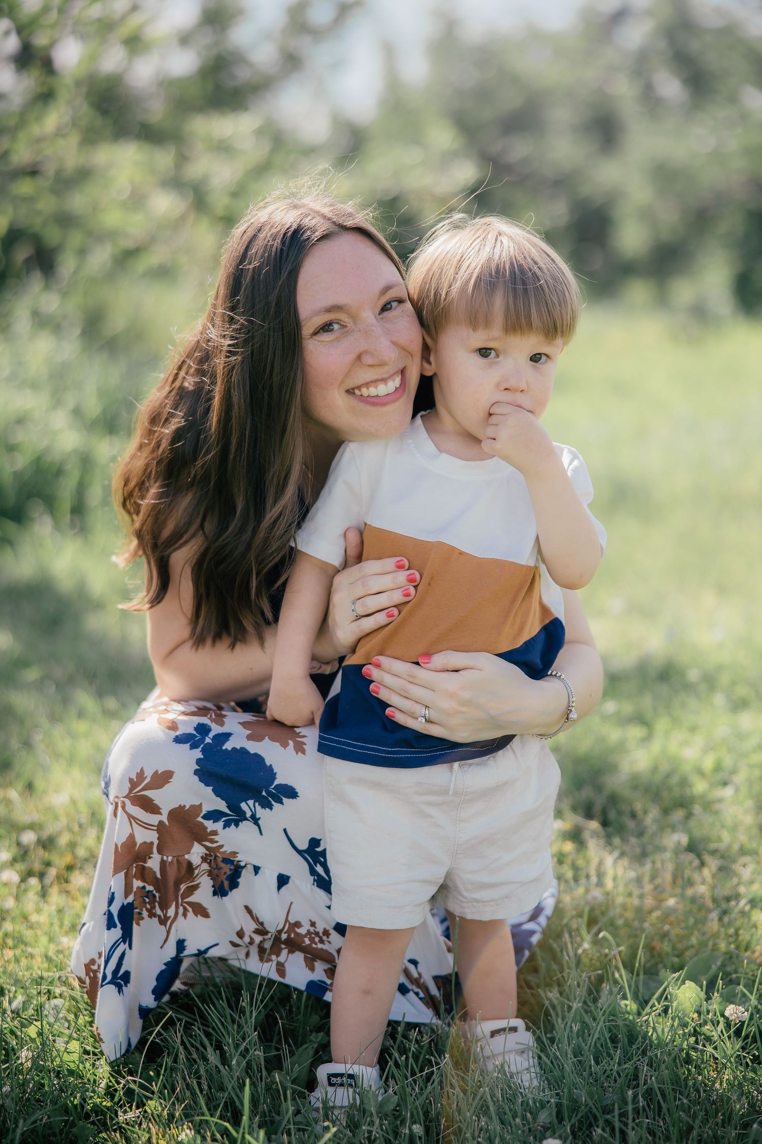 Smiling mother crouching in sunny green field hugging her young toddler son in casual summer clothes.