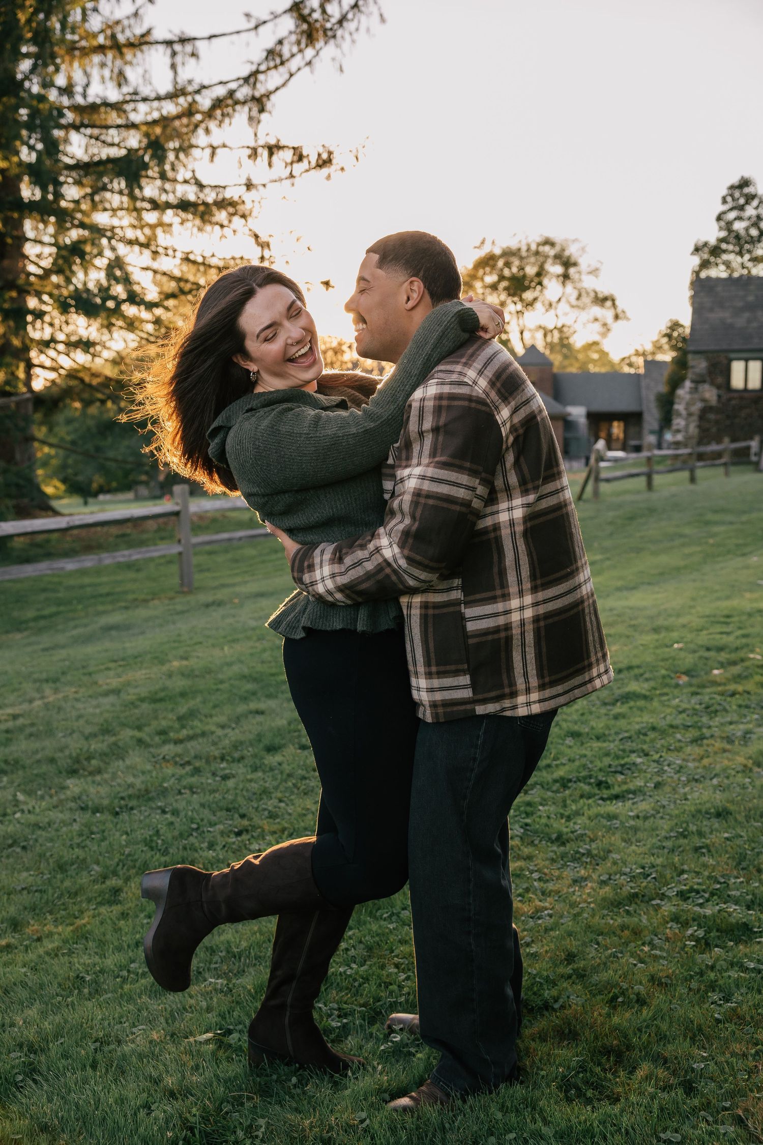 A joyful couple embraces and laughs together on a green lawn at sunset, wearing cozy fall outfits near a rustic stone building.