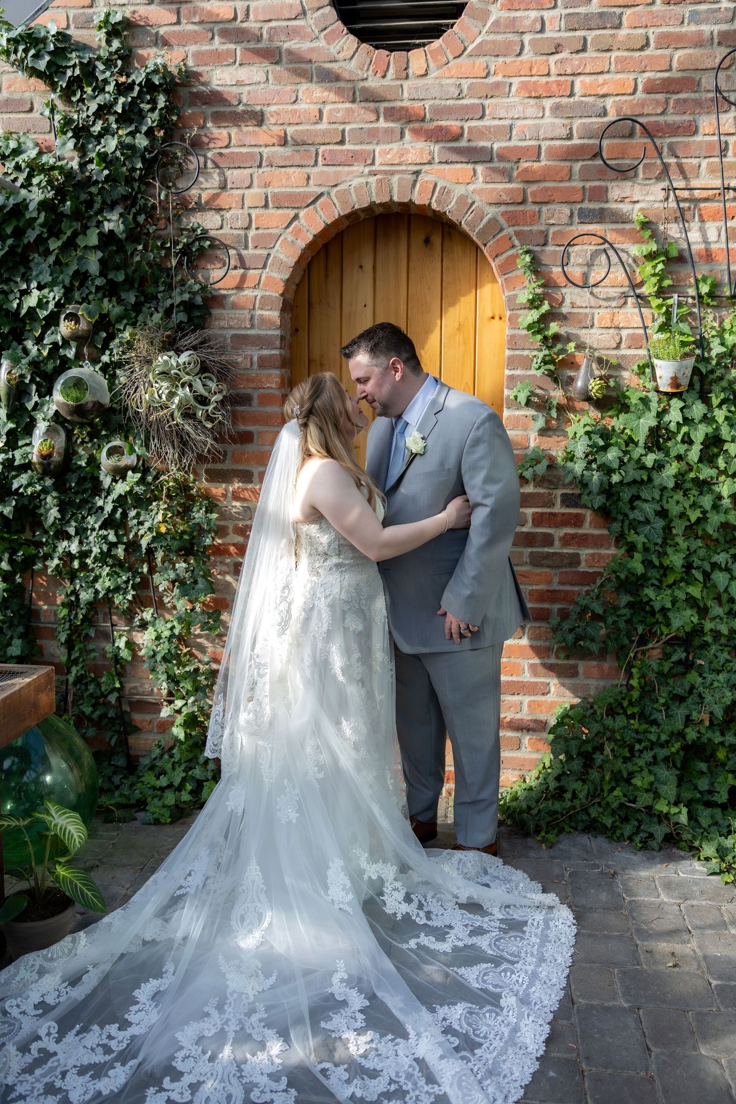 Bride and groom share a romantic moment in front of a rustic brick wall with arched wooden door and ivy.