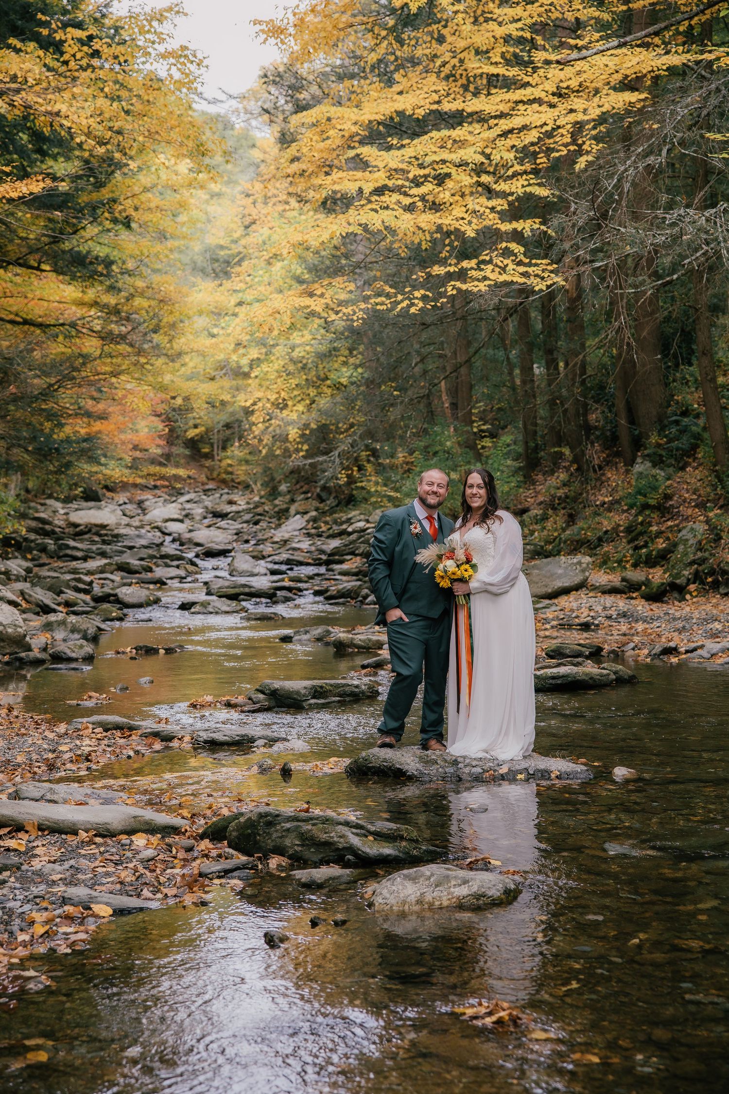Couple in wedding attire standing on rocks by a stream surrounded by vibrant autumn foliage in a forest.