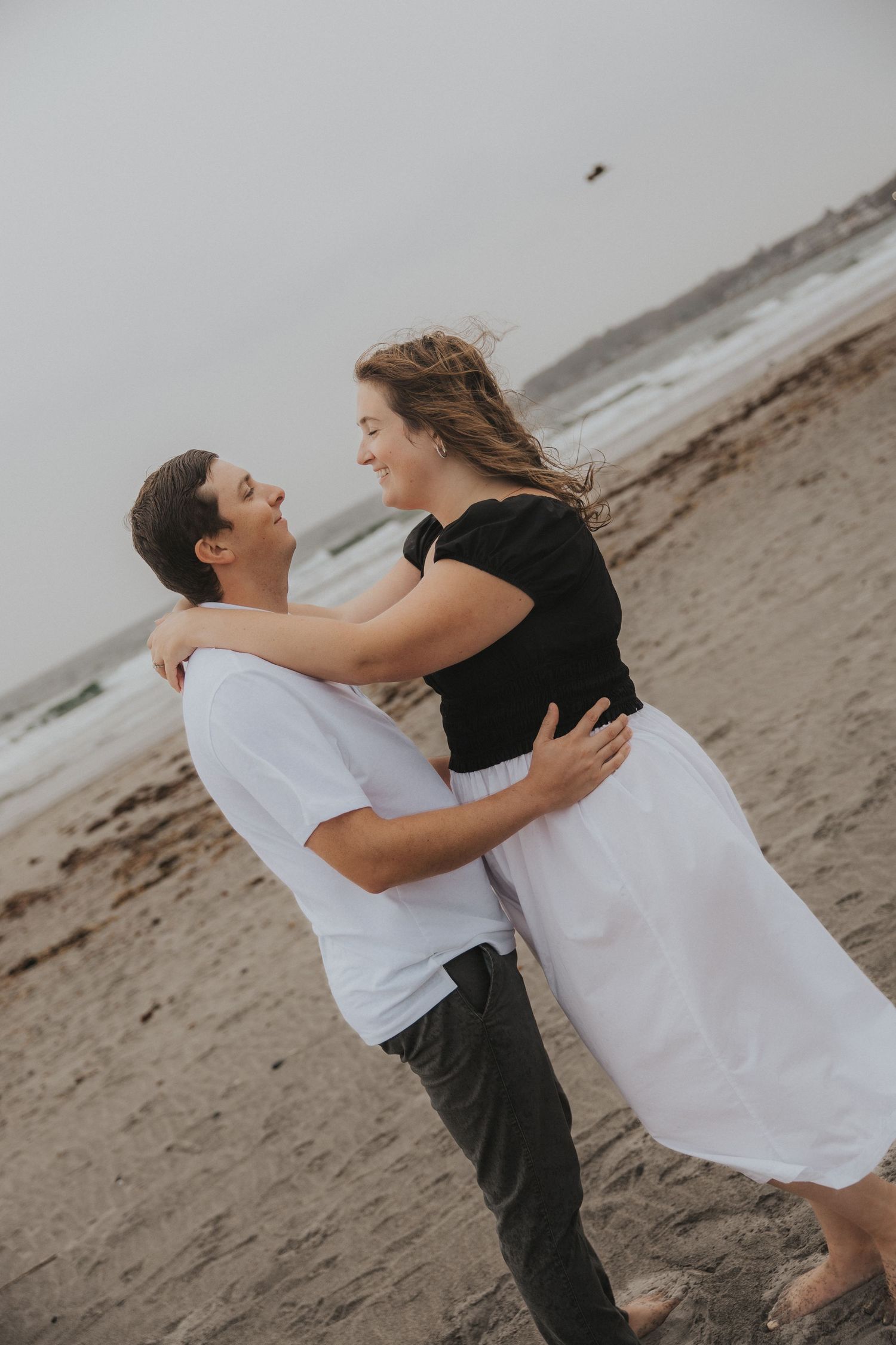 A smiling couple embraces on a windy, overcast beach, the woman in a black top and white skirt, the man in a white shirt.