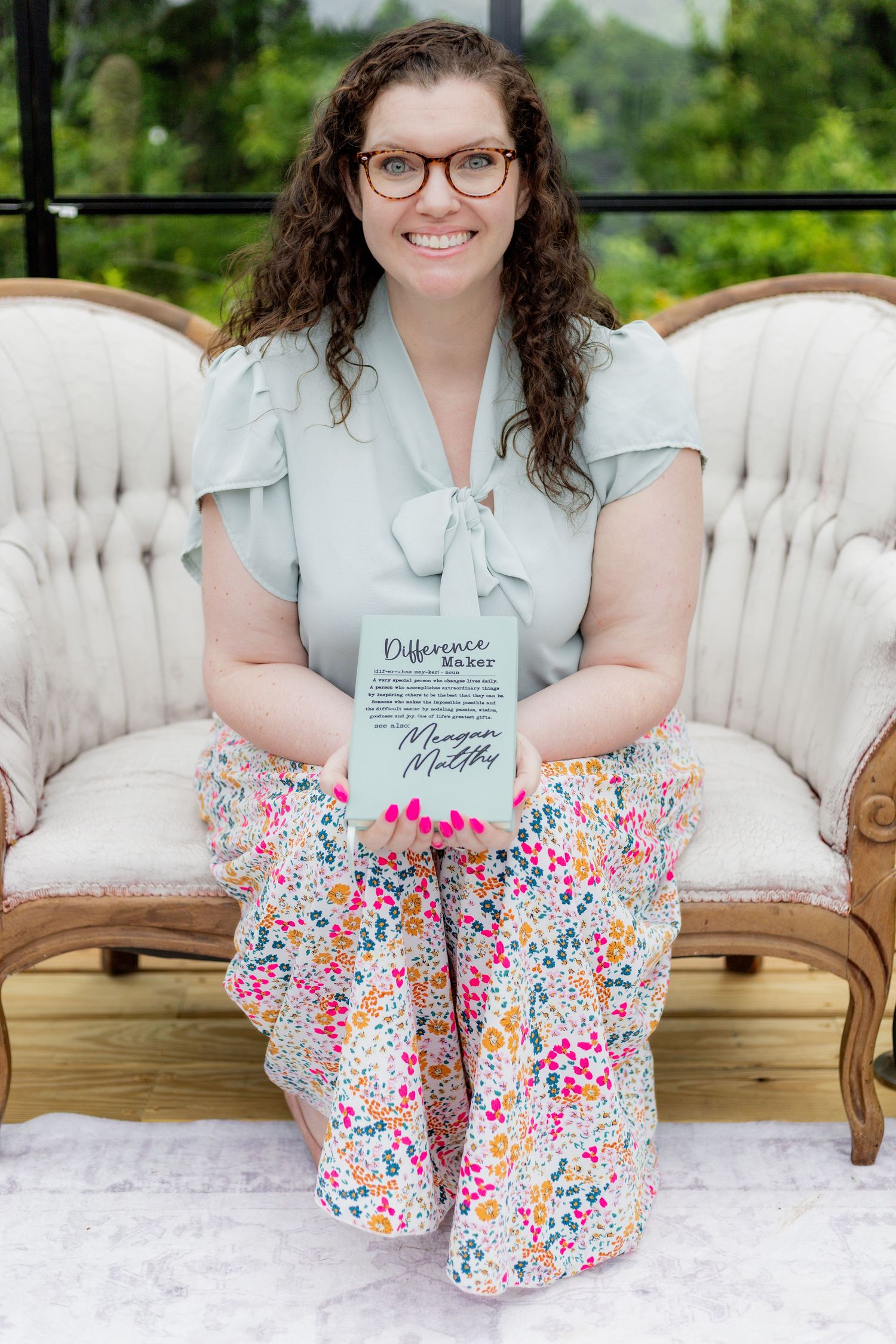 Person in light blue shirt and floral pants poses on vintage sofa with decorative pillows.