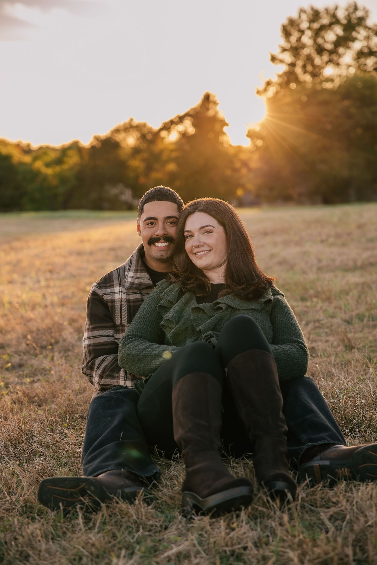 Couple sitting together in a golden sunset field, man in plaid jacket and woman in green coat, smiling warmly.