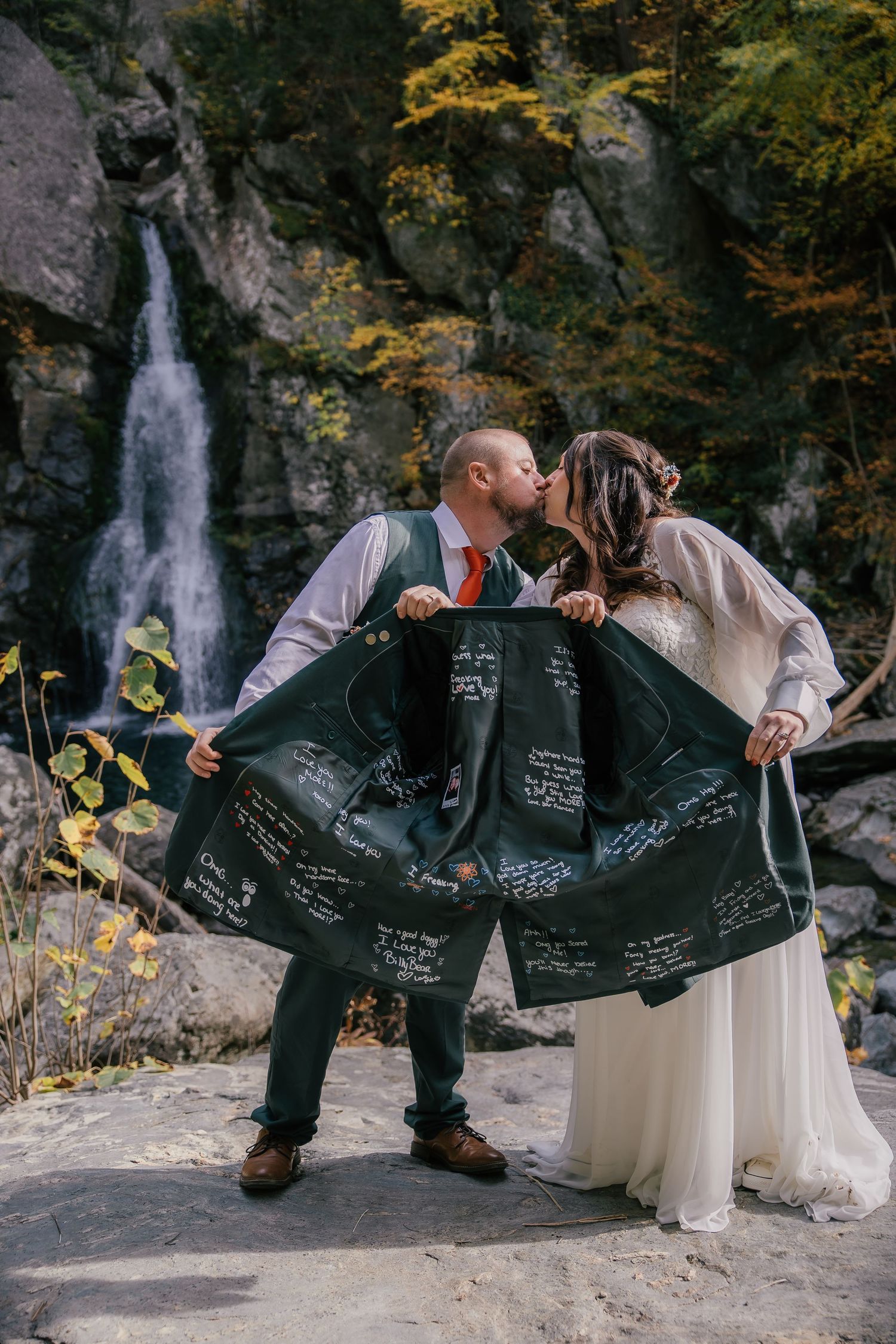 Bride and groom kissing outdoors near a waterfall, displaying a jacket signed with wedding guest messages.
