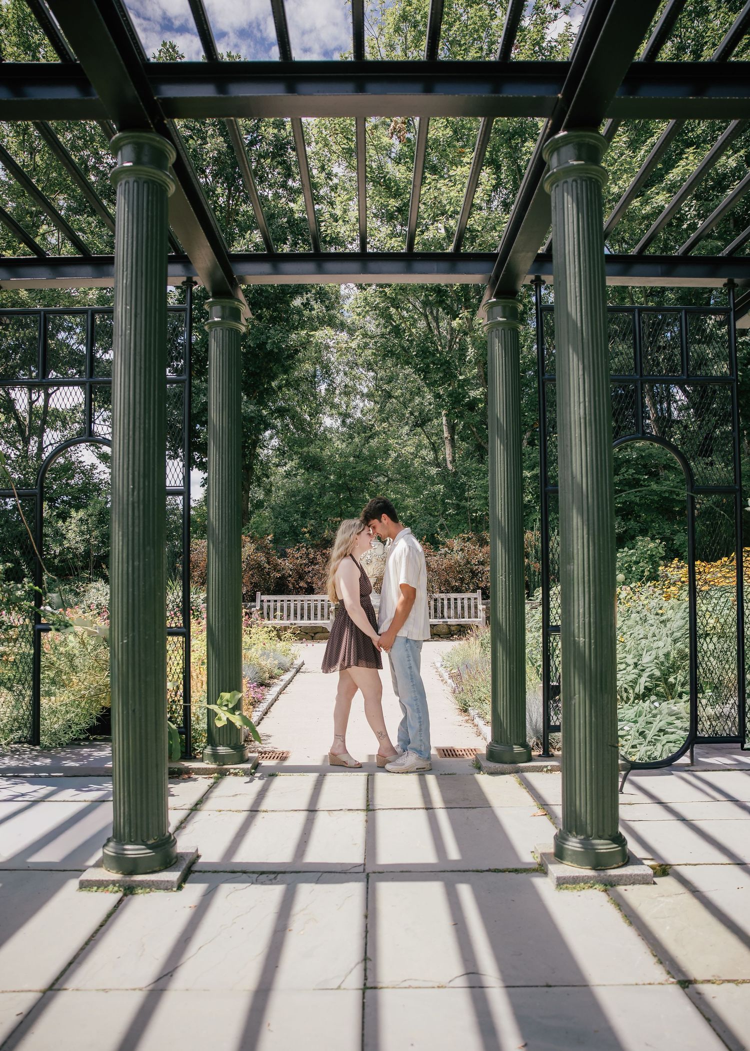 Couple sharing a romantic kiss under a green pergola in a lush garden setting with dappled sunlight.