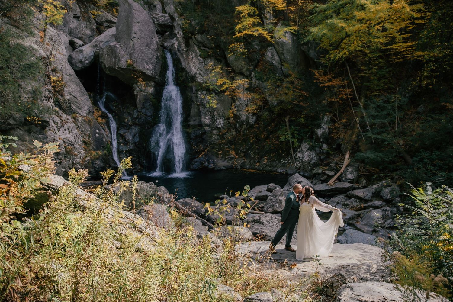 Bride and groom dance on rocky terrain before a waterfall surrounded by autumn foliage in a forest setting.