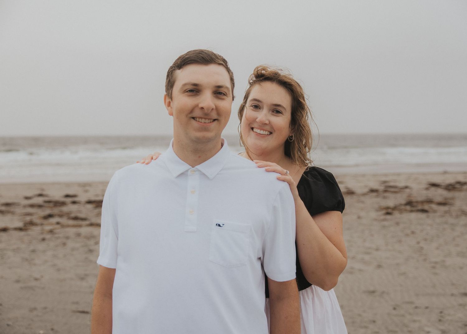 Smiling couple posing together on a sandy beach, the woman hugging the man from behind, with ocean waves and an overcast sky behind them.