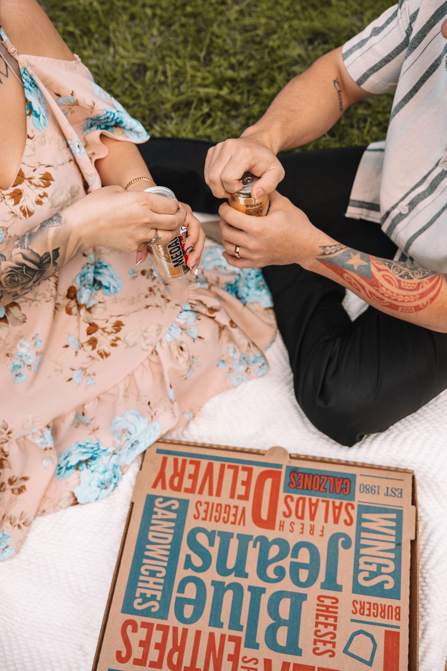 Couple enjoying a picnic with Blue Jeans pizza box, canned drinks, sitting on white blanket outdoors.
