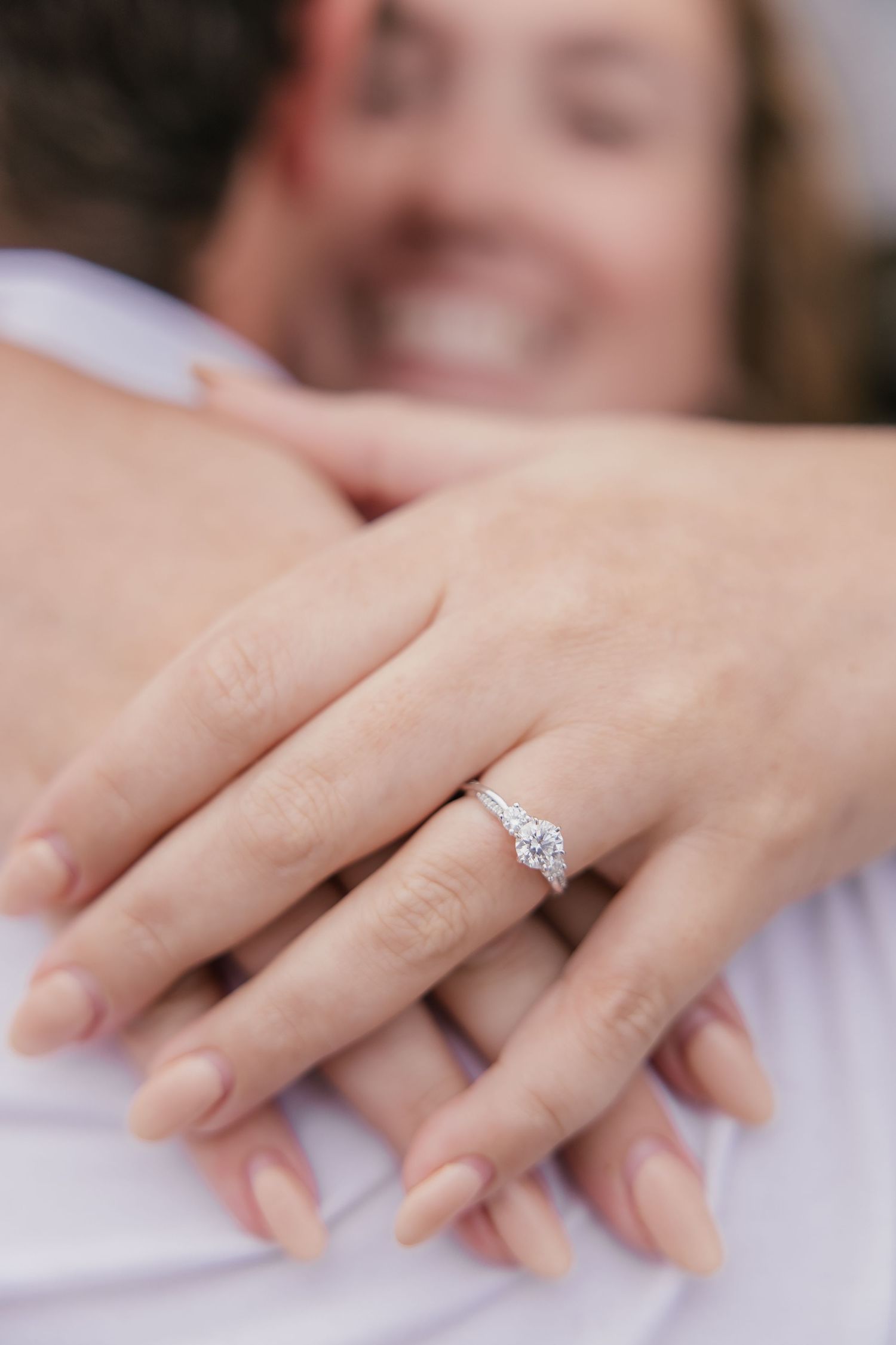 Close-up of a diamond solitaire engagement ring on a woman's hand as she embraces her smiling partner.