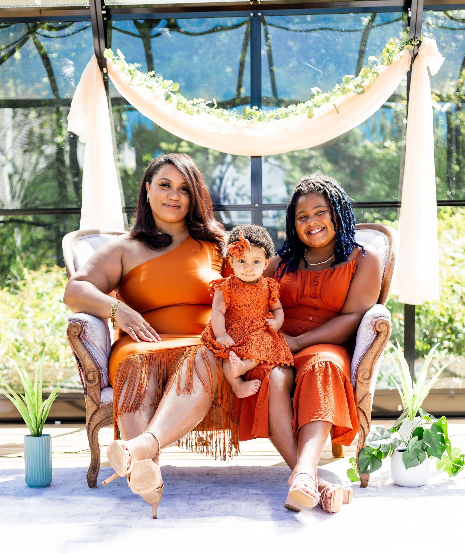Three people in orange outfits sit on a swing bench under a floral arch.