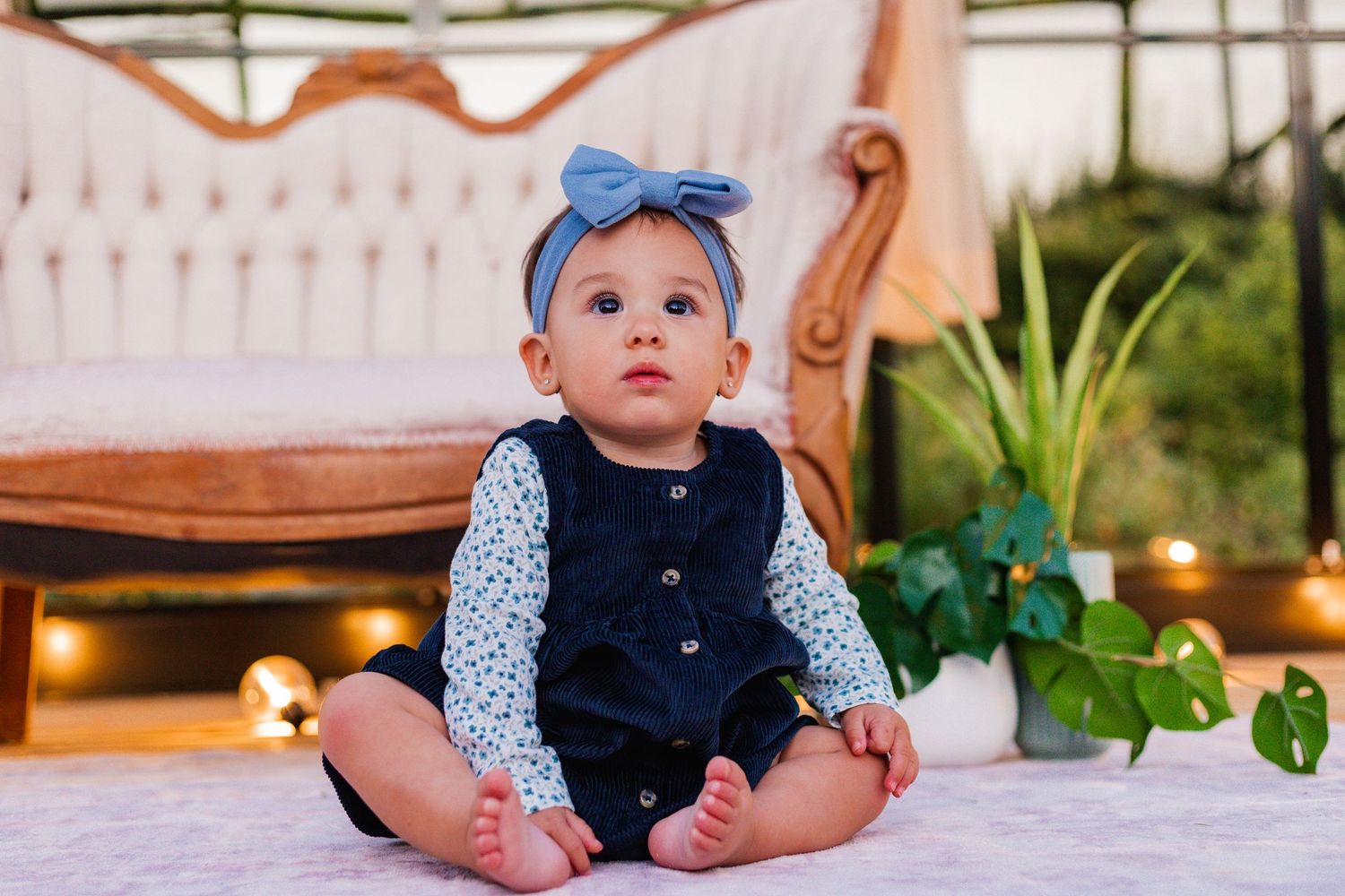 A baby in a navy outfit and blue bow headband sits on a white surface near indoor plants.