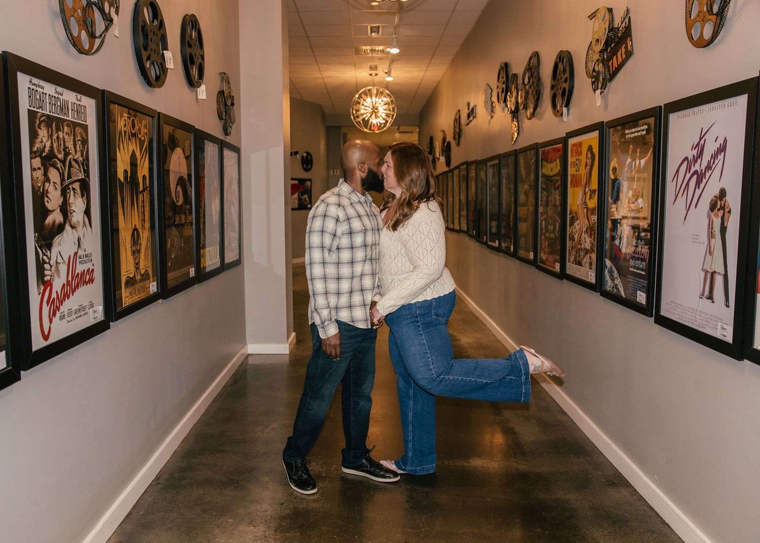 Couple sharing a romantic moment in a hallway lined with framed movie posters and vinyl records on white walls.