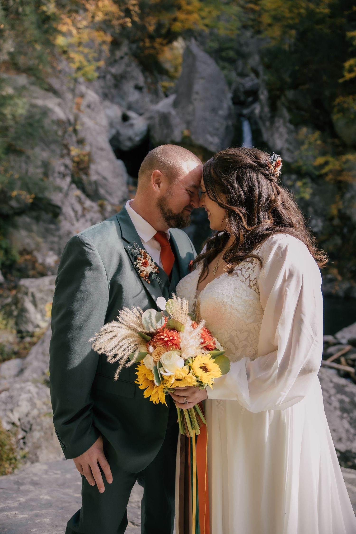 A bride and groom touch foreheads tenderly outdoors, holding a colorful fall bouquet with sunflowers near a rocky waterfall.