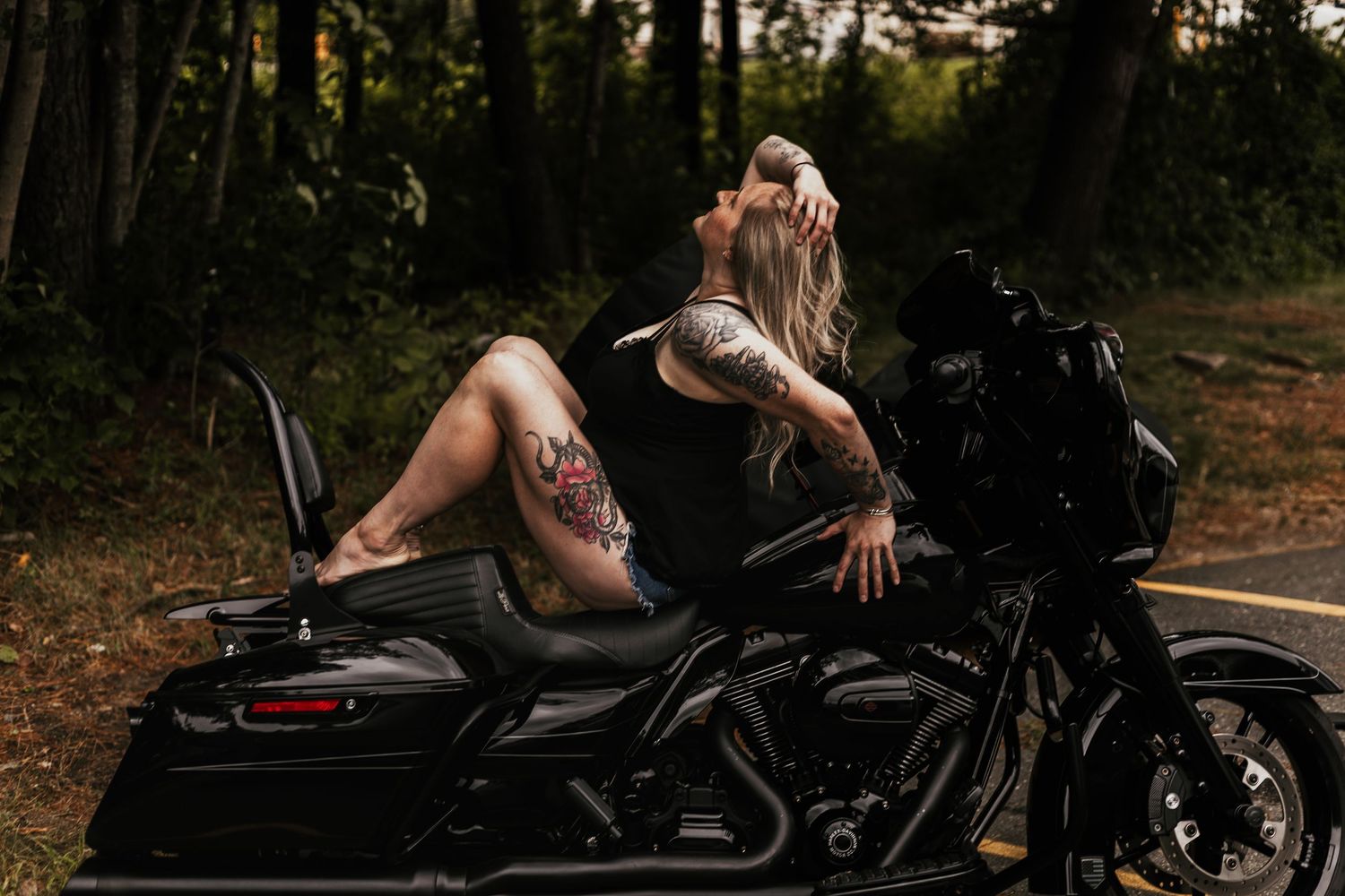 Woman in black poses on a parked motorcycle in a forest setting.