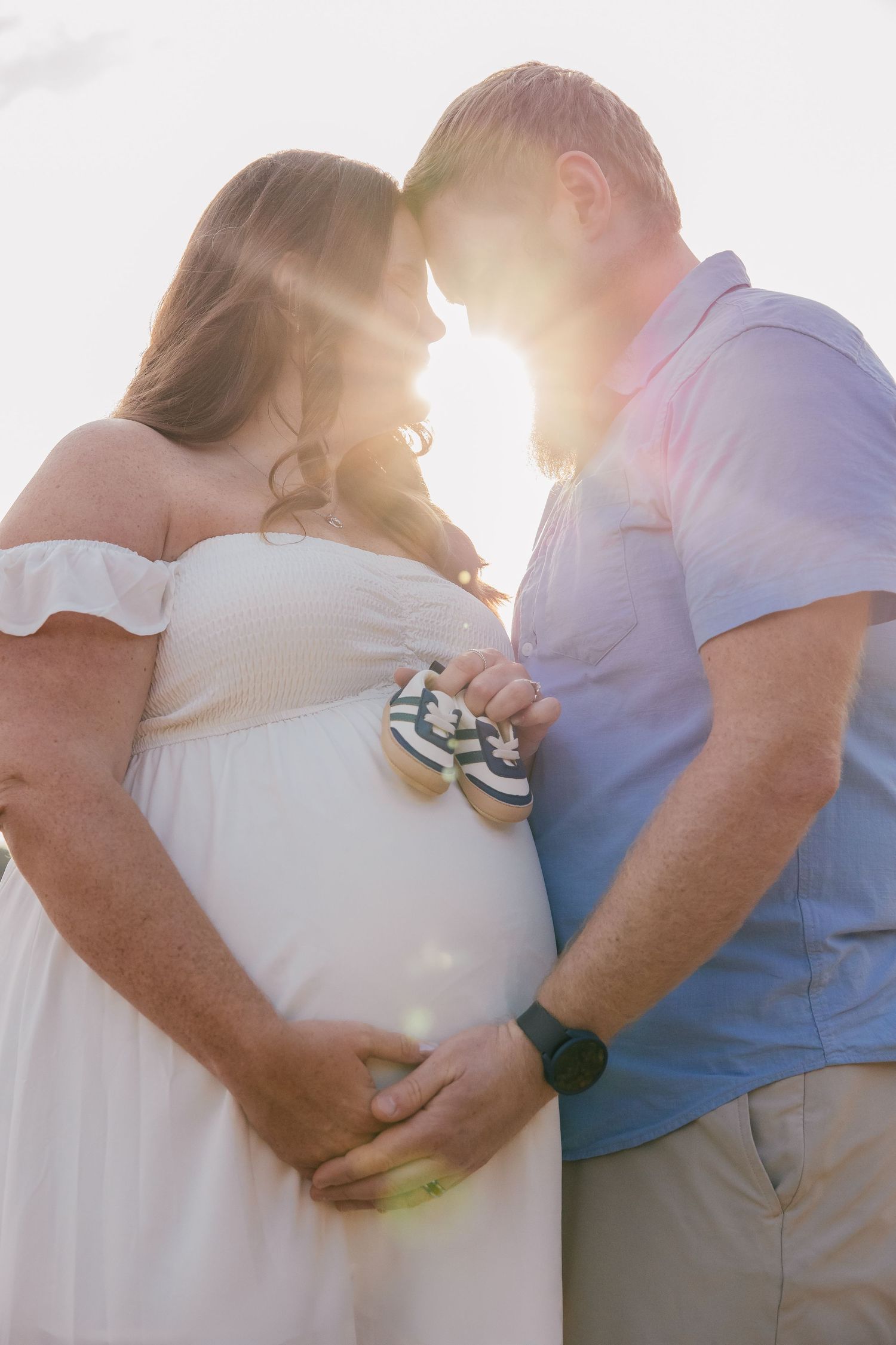 Pregnant couple holding tiny baby shoes against glowing sunset, man and woman touching foreheads tenderly.