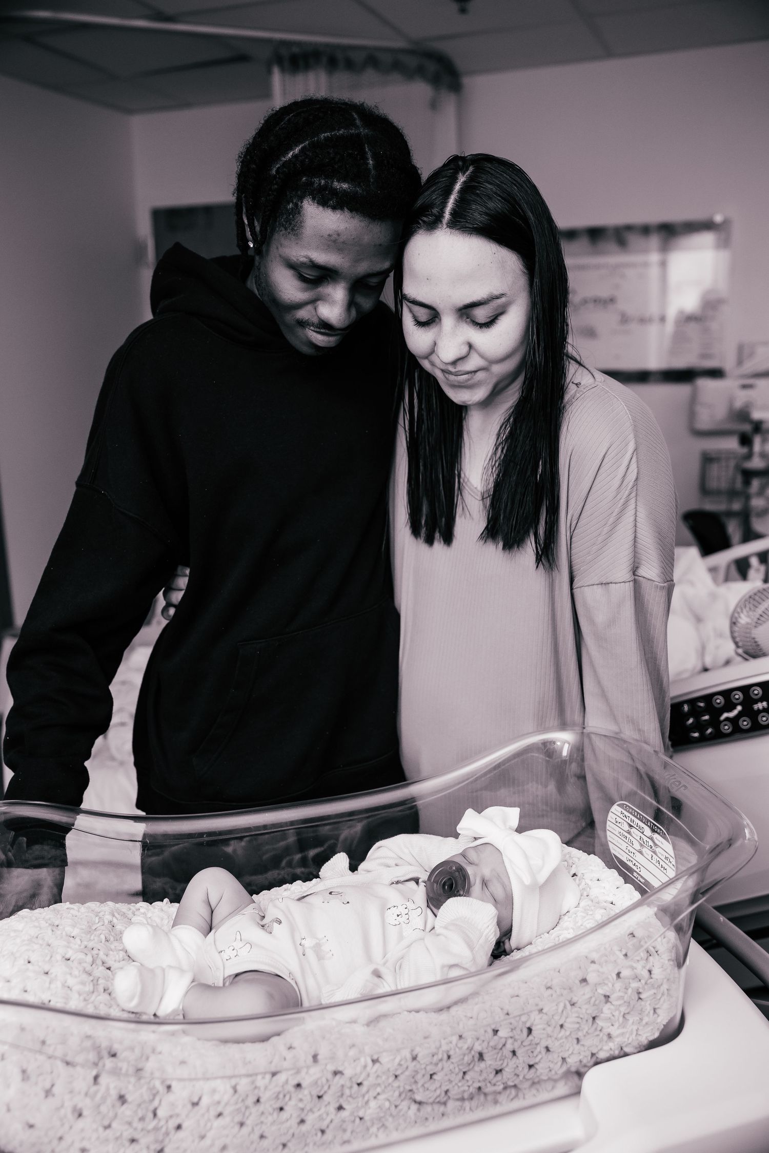 Black and white photo of new parents lovingly gazing at their newborn baby in a hospital bassinet.