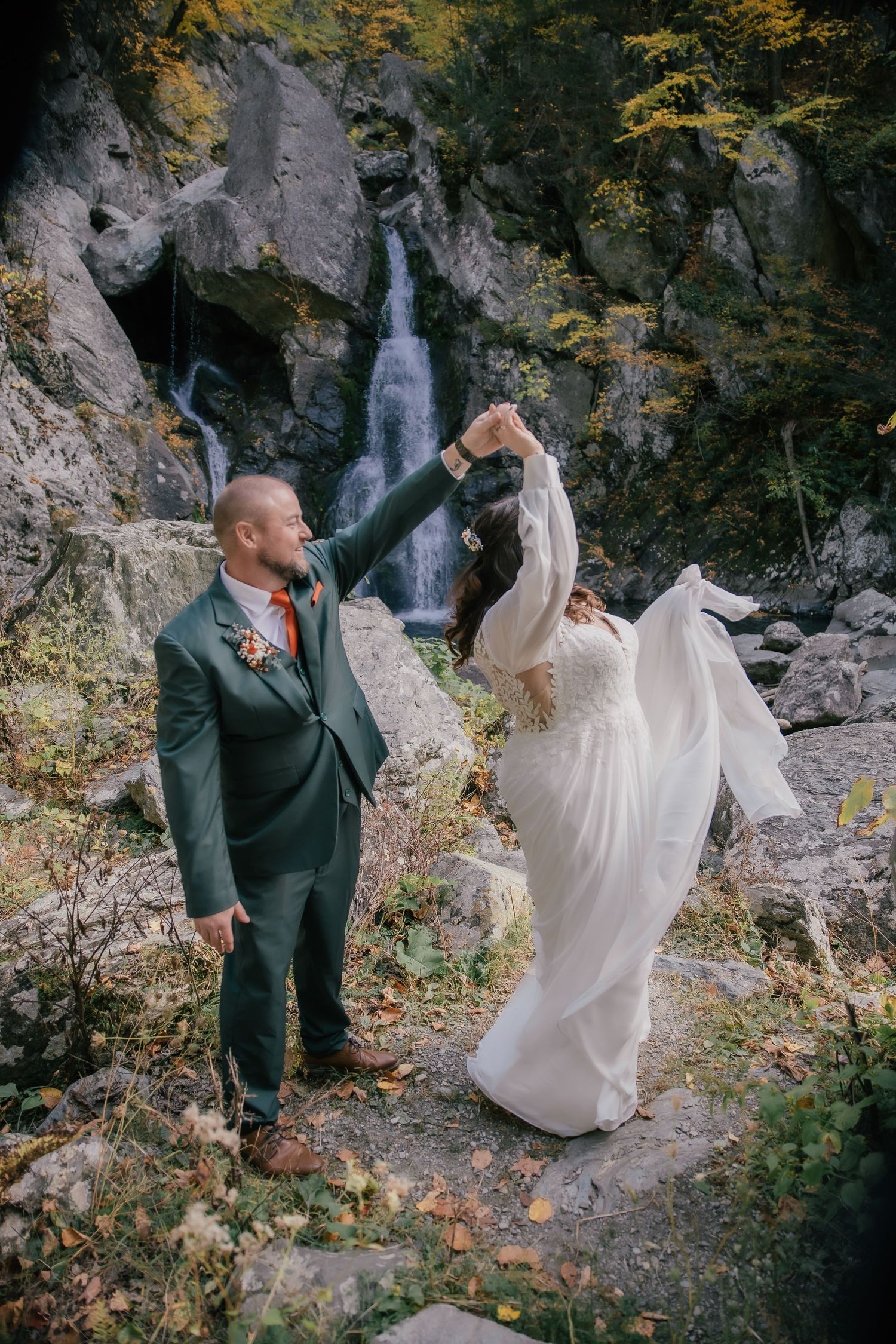 A couple dances in front of a waterfall during their outdoor fall wedding, bride in white gown, groom in green suit.