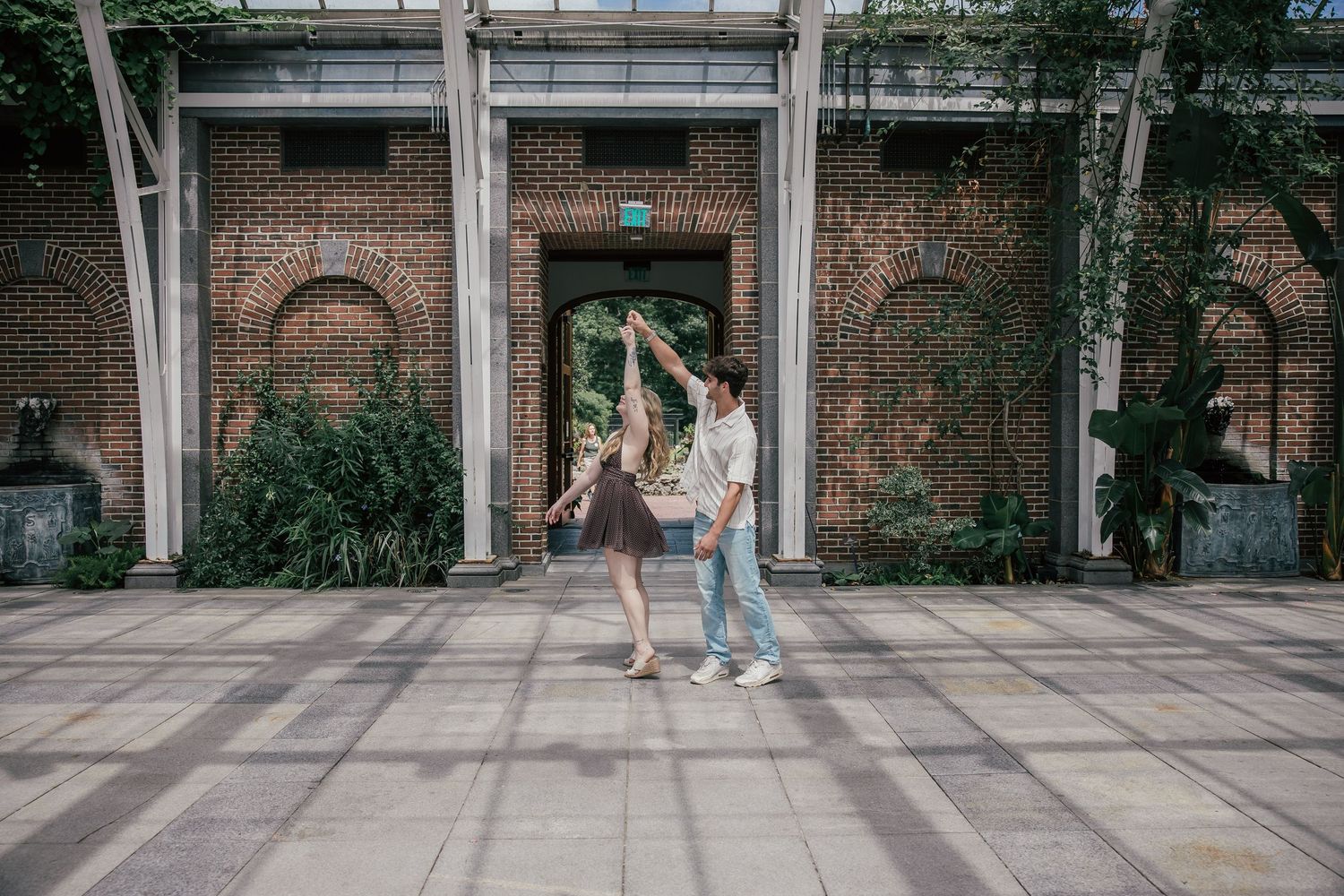 Couple dancing together in front of a brick archway building surrounded by lush greenery on a tiled plaza.