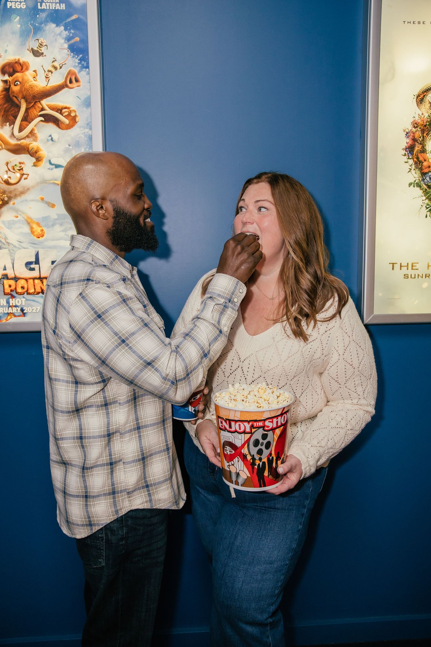 A man in a plaid shirt playfully feeds popcorn to a smiling woman holding a striped popcorn bucket in a movie theater lobby.