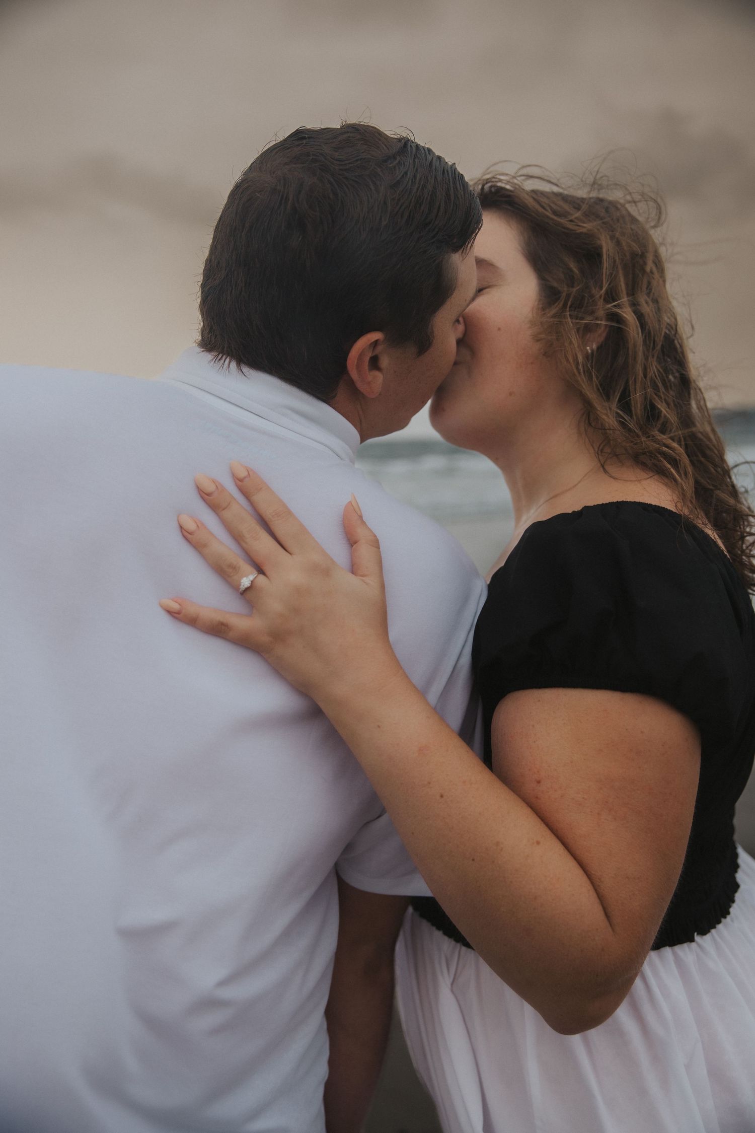 Couple kissing on a windswept beach at sunset, the woman wearing a ring and black top with a white skirt, hand resting on his shoulder.