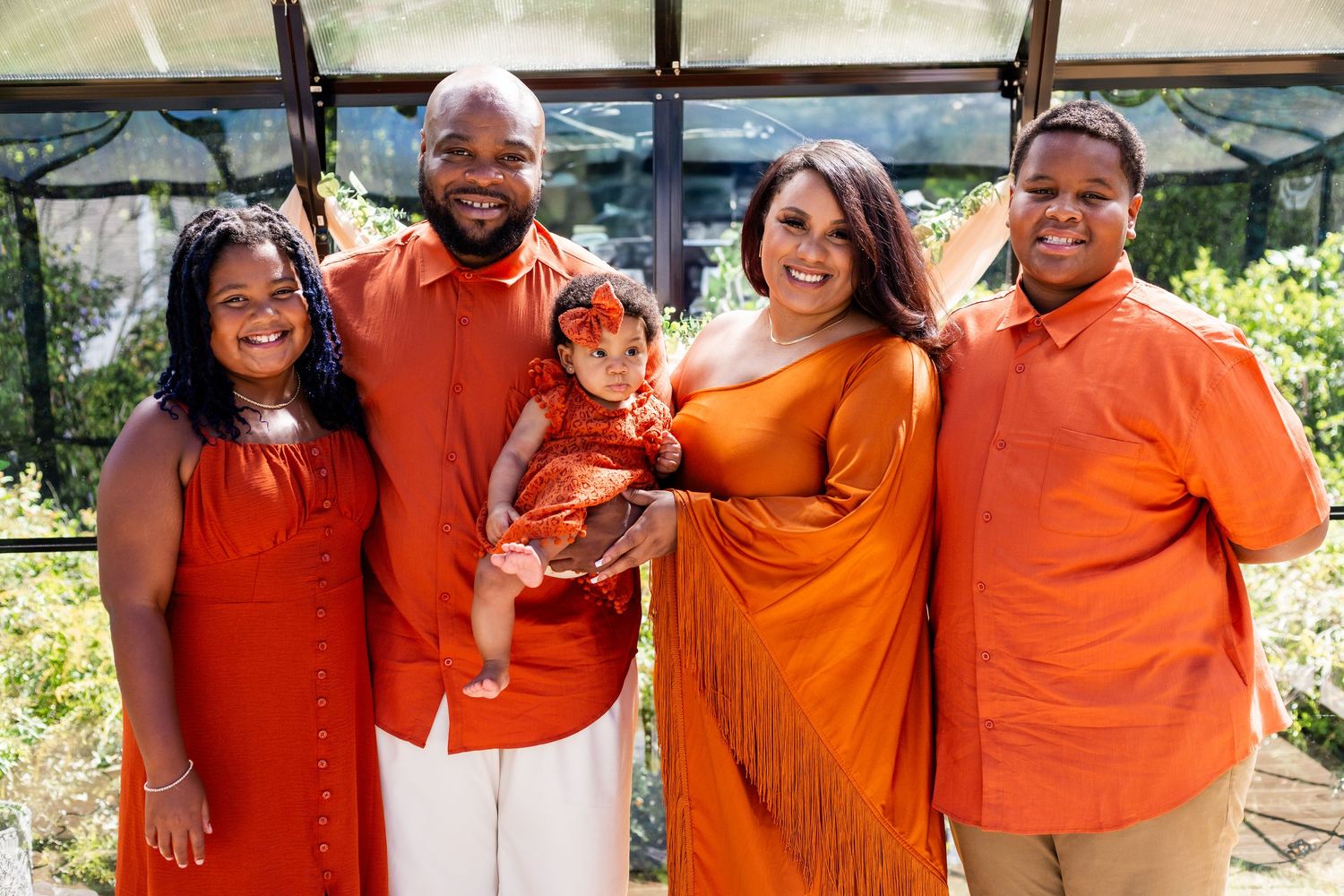 A group in coordinated orange and white outfits poses together in a greenhouse setting.