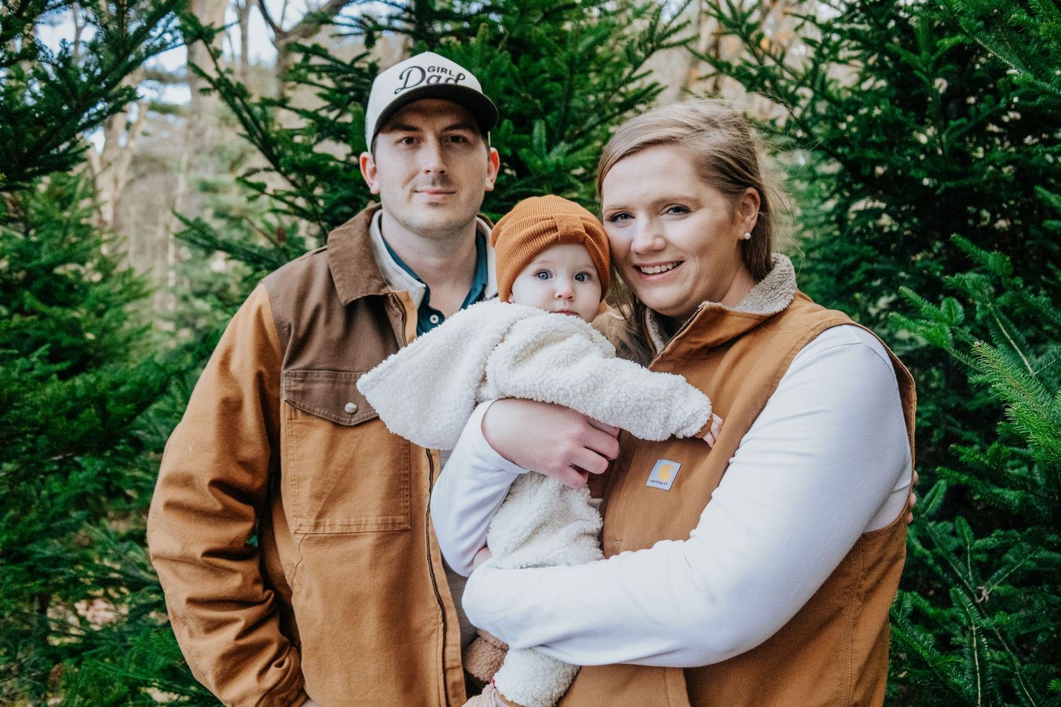 Family wearing earth-toned clothing embracing outdoors among evergreen trees.